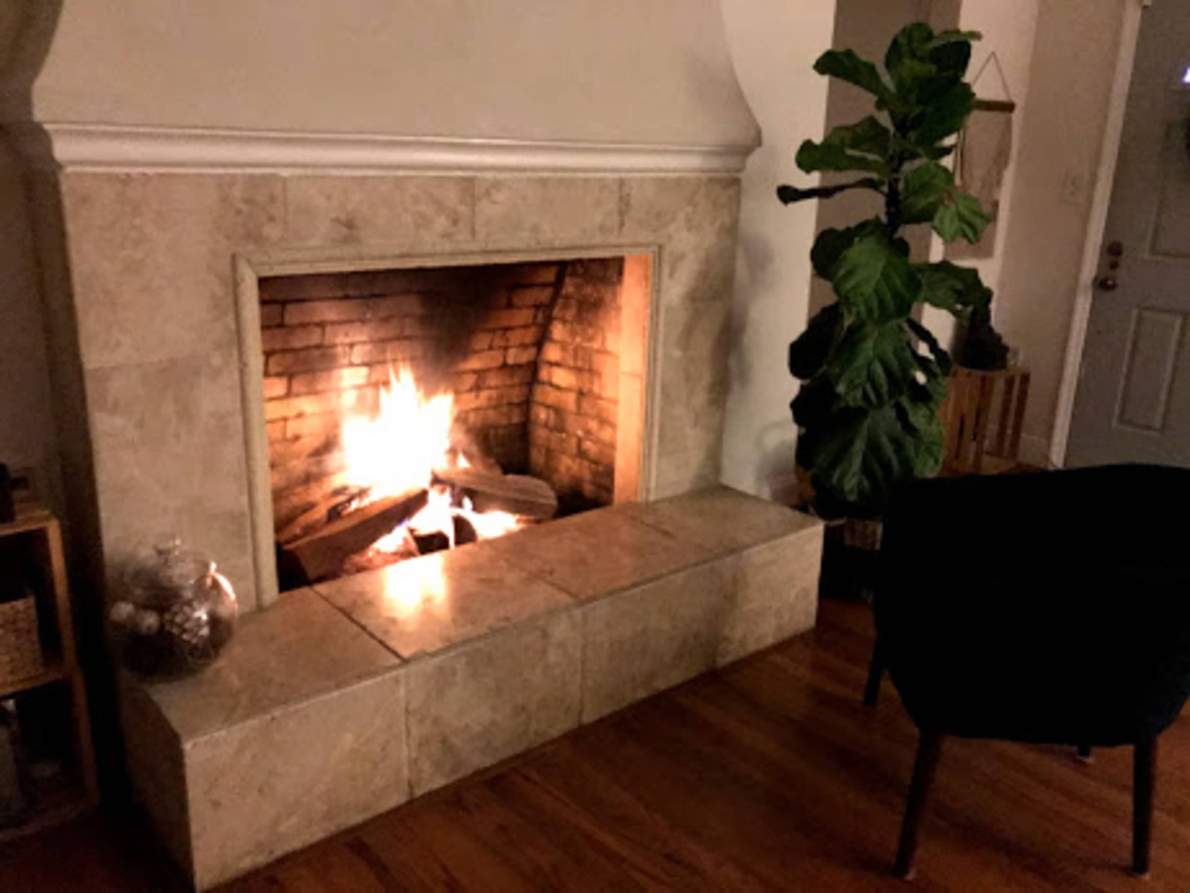 A fireplace with a lit fire, surrounded by a stone mantle, a potted plant, and a chair in a room with wooden flooring.