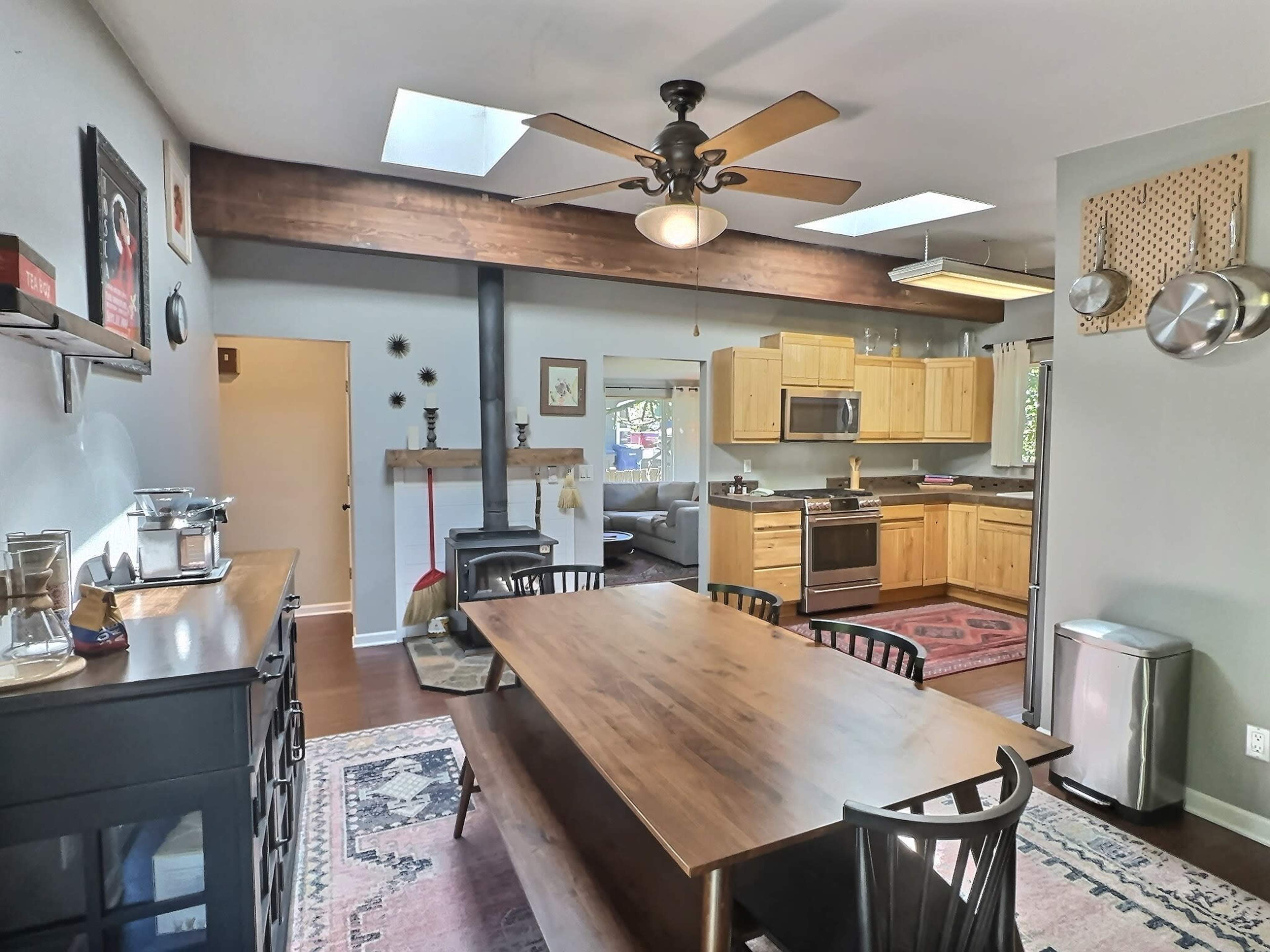 The image depicts a modern kitchen and dining area featuring a wooden dining table, black chairs, stainless steel appliances, and a light ceiling fan, with natural light streaming in through skylights.