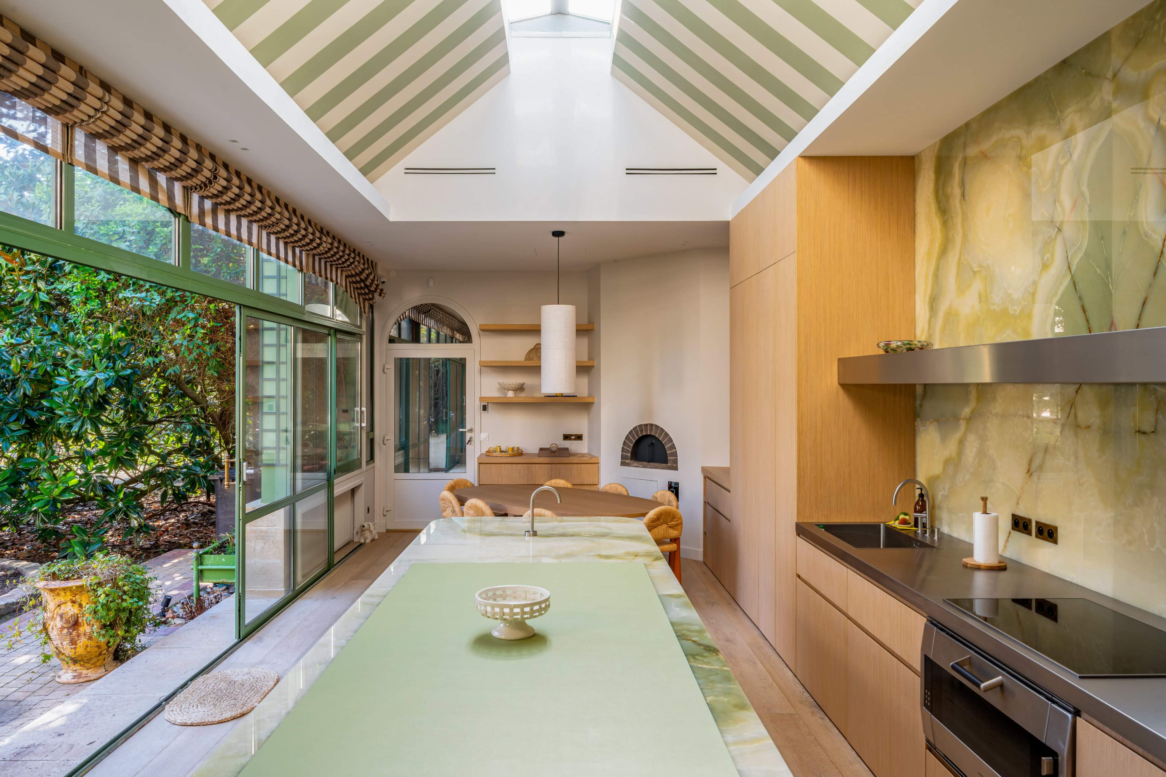 The image shows a modern kitchen with a green marble island, wooden cabinetry, and large windows that provide natural light.