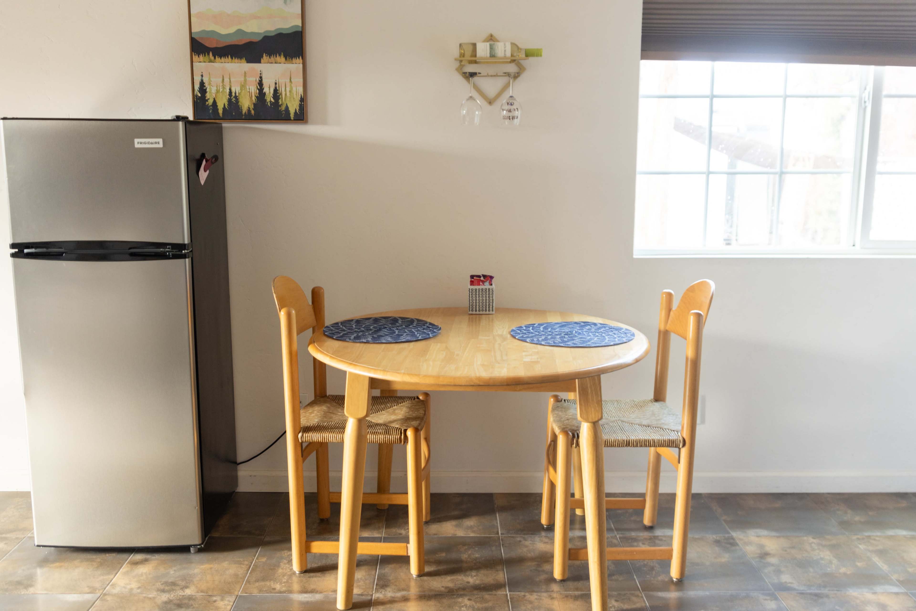A small round wooden table with two wooden chairs is positioned near a gray refrigerator in a brightly lit kitchen.
