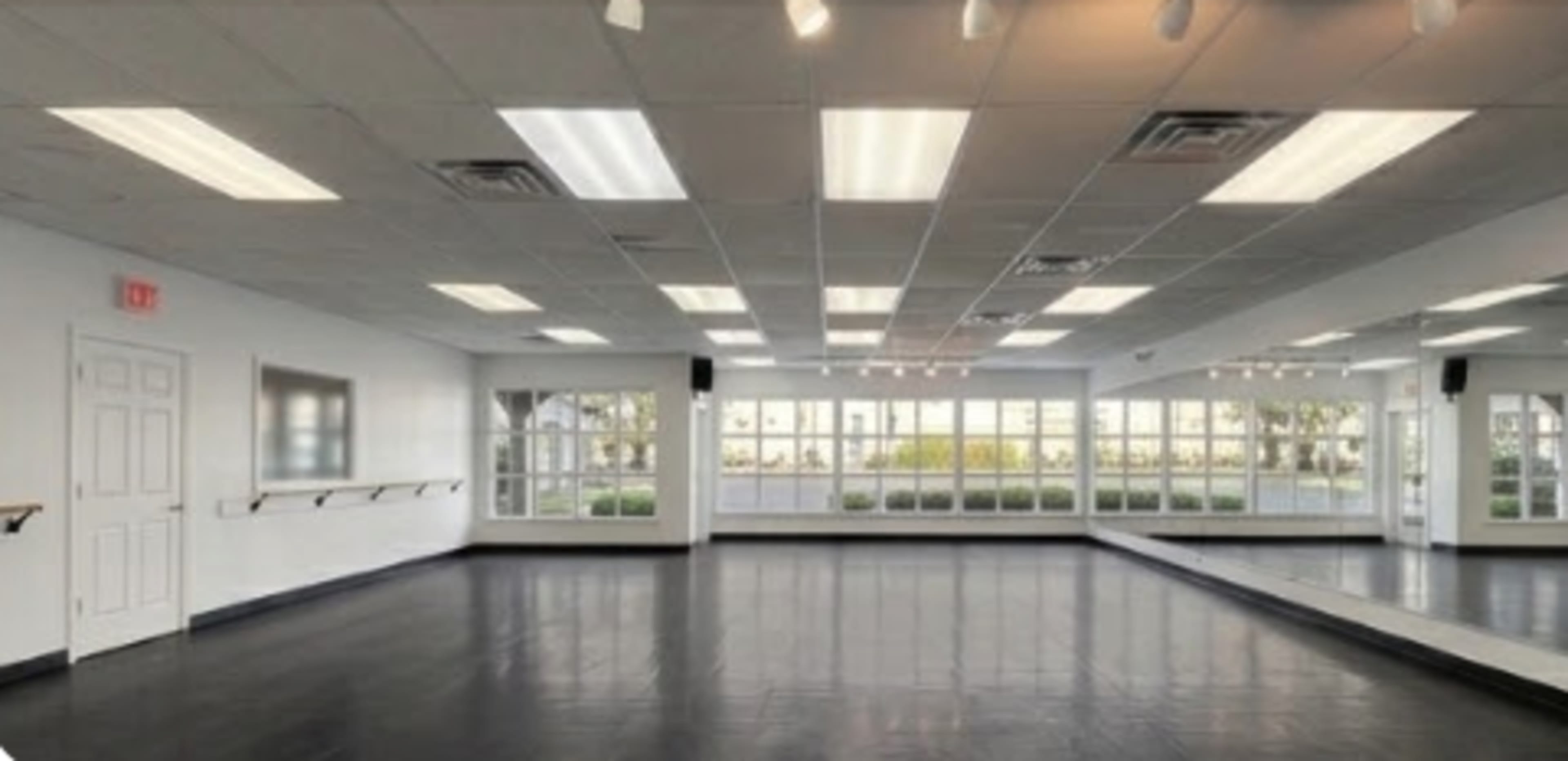 The image shows a spacious dance studio with wood floors, large windows, and mirrored walls under bright lights.