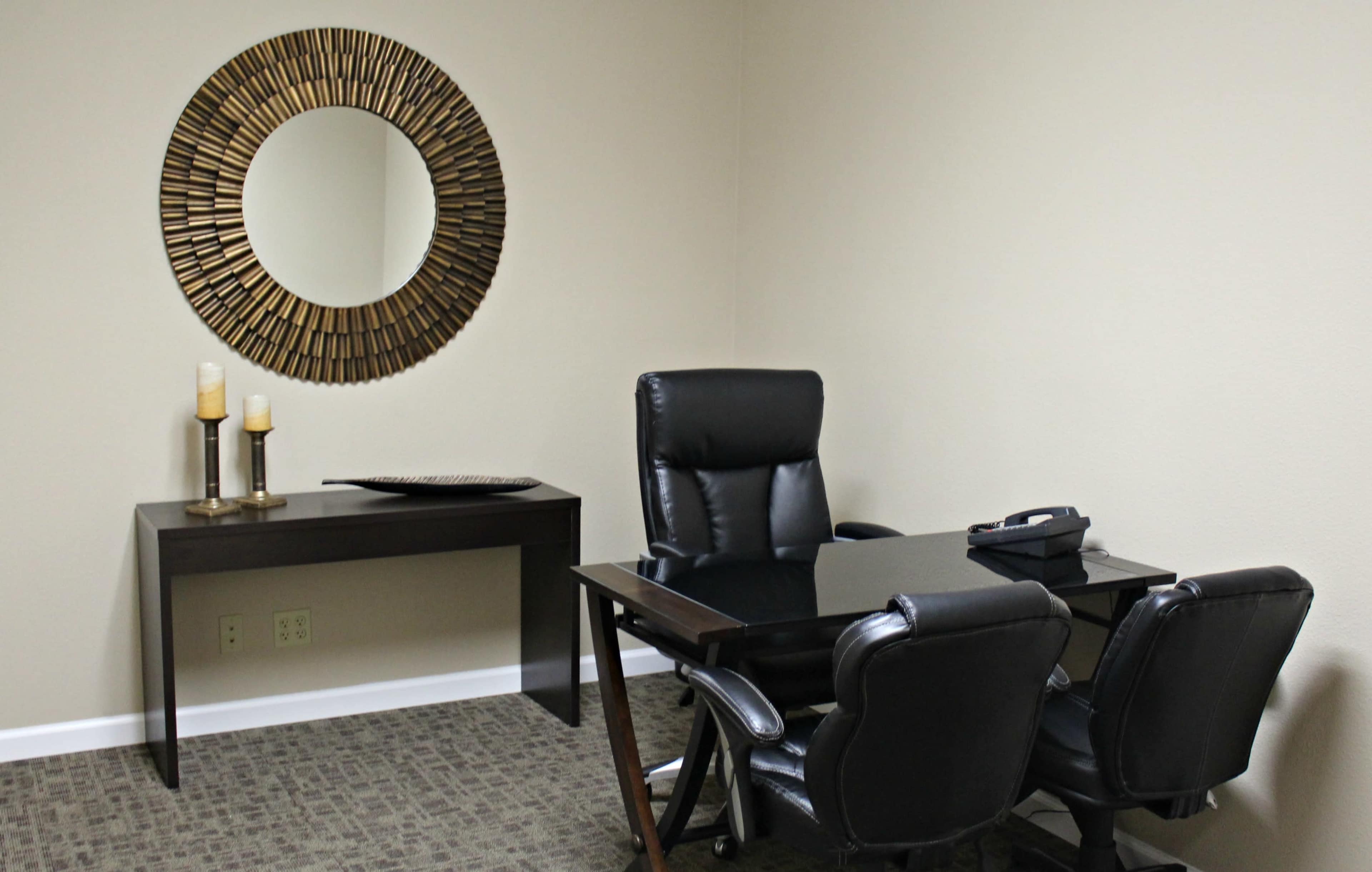 A minimalist office features a black desk, two office chairs, a telephone, a round mirror, and candles on a side table against a beige wall.