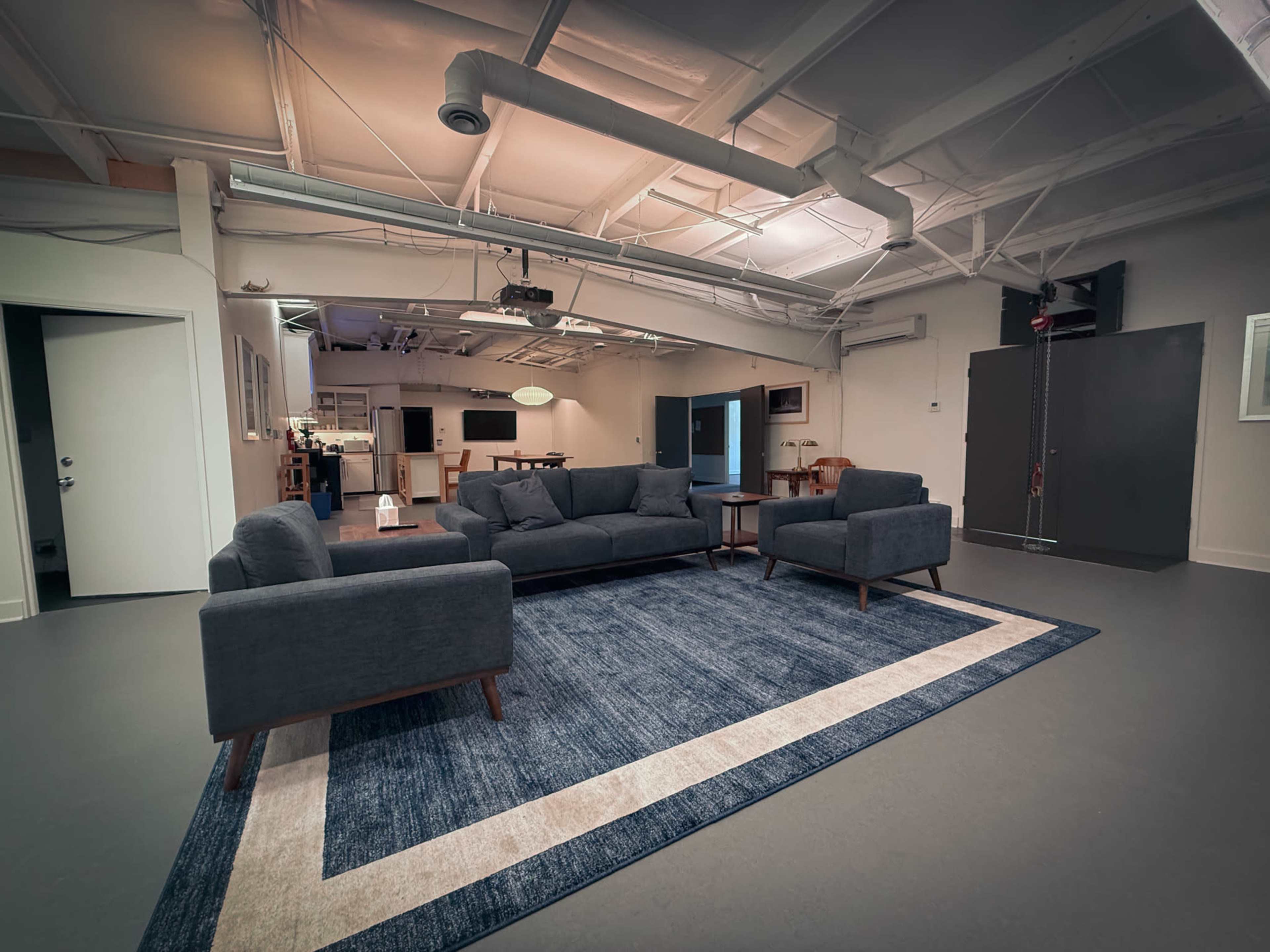 The image shows a spacious, modern room with a gray sofa set arranged on a large area rug, featuring a minimalist design and neutral colors.