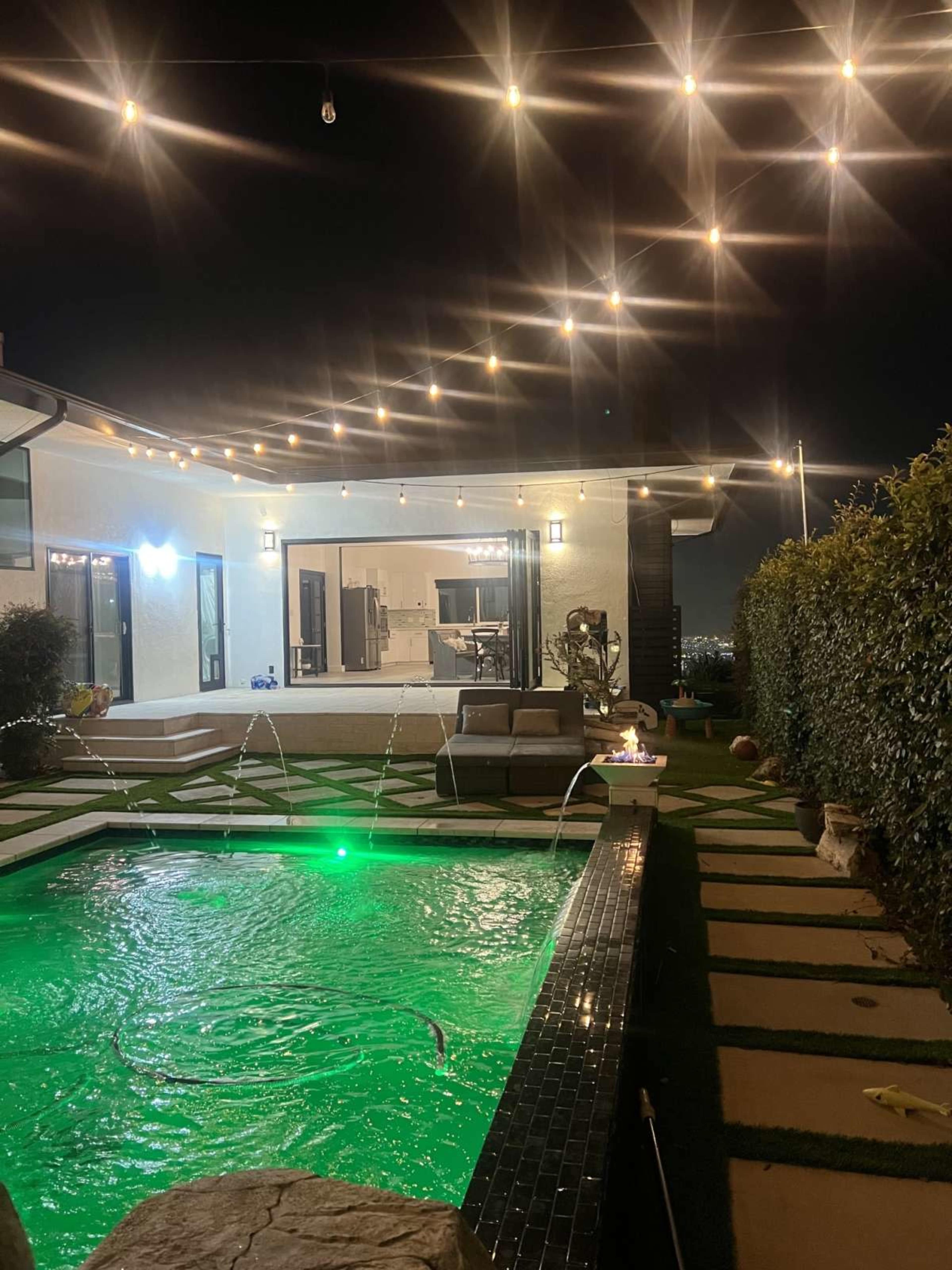 A modern backyard features a glowing green pool with water fountains, surrounded by patio furniture and string lights overhead, against a backdrop of a well-lit, contemporary home.
