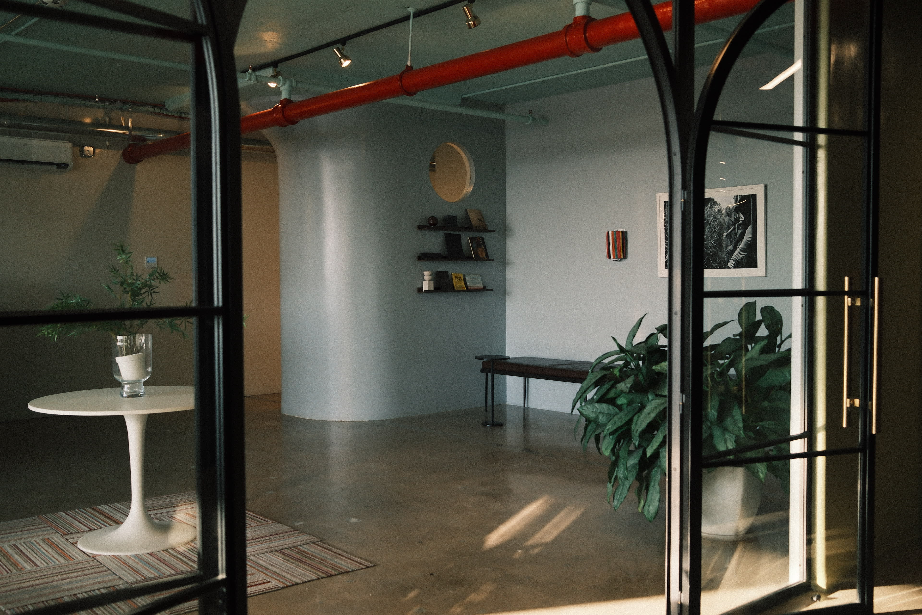 The image shows a modern interior space with a round table and plants, featuring a minimalist design with shelves on the wall and large glass doors.
