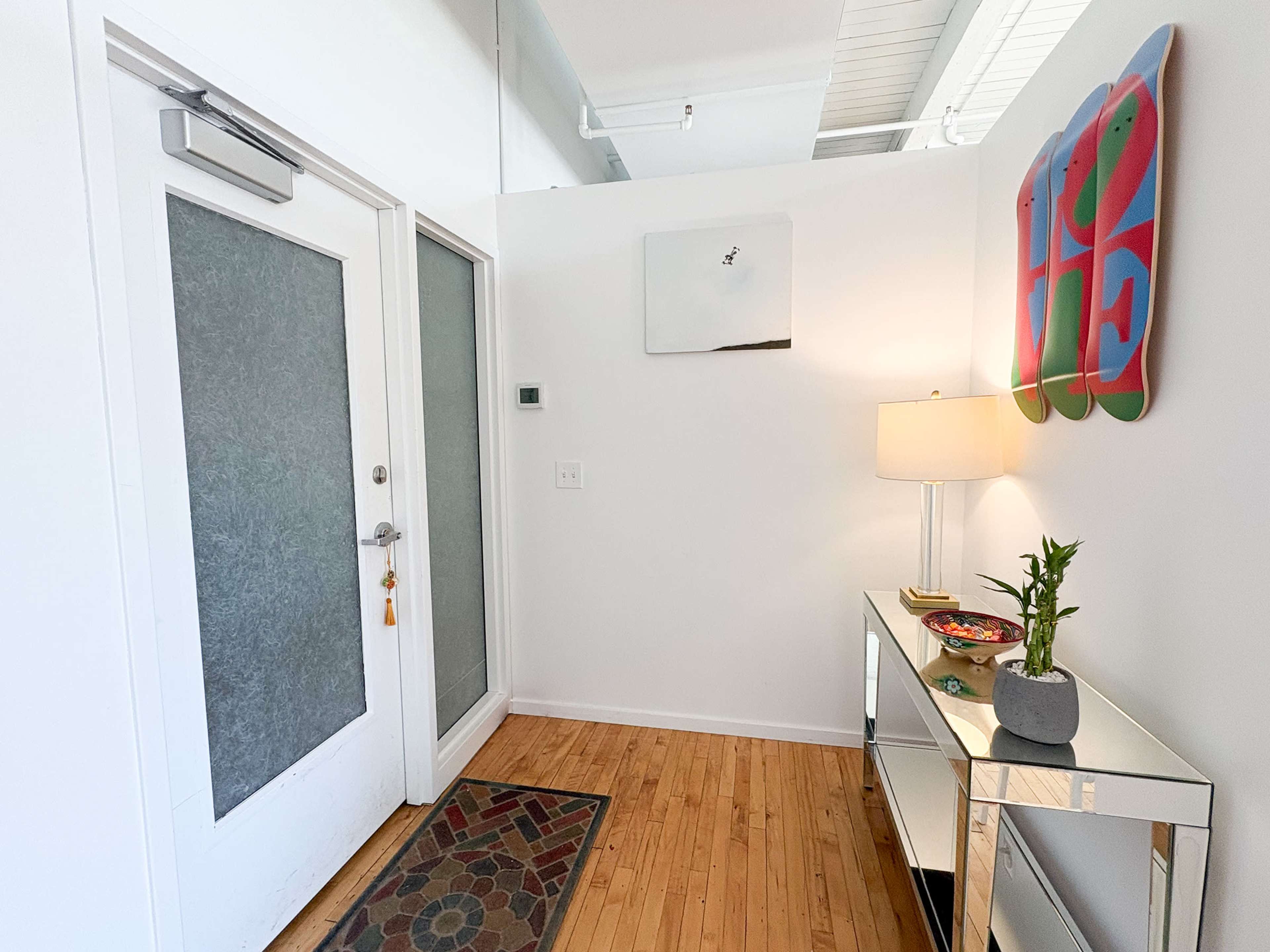 The image depicts a modern entryway with a glass-paneled door, a contemporary lamp on a mirrored console table, and an artistic wall decoration.