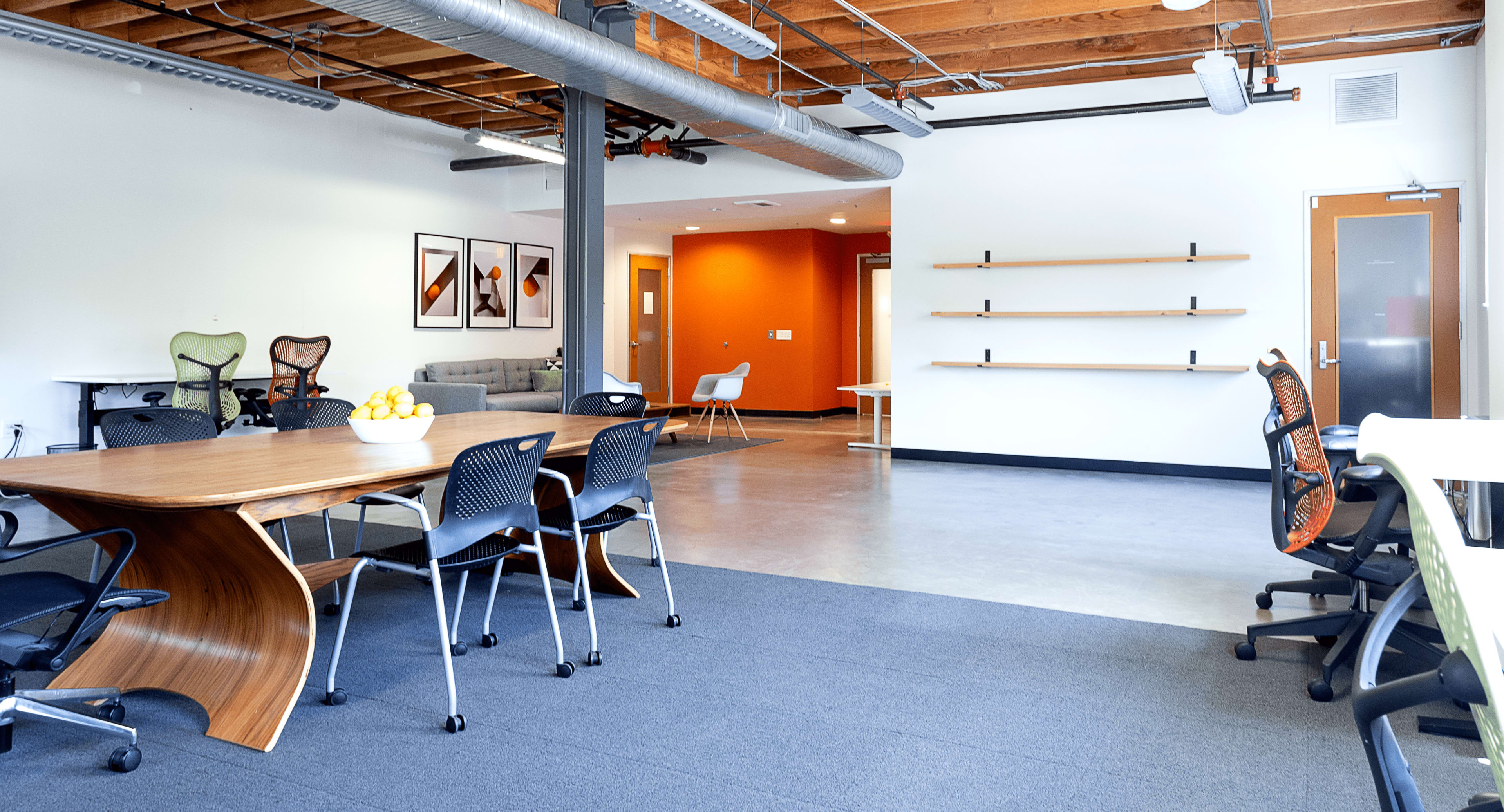 A modern office space featuring a wooden table with chairs, a seating area, and minimalist shelves against a backdrop of light and orange walls.