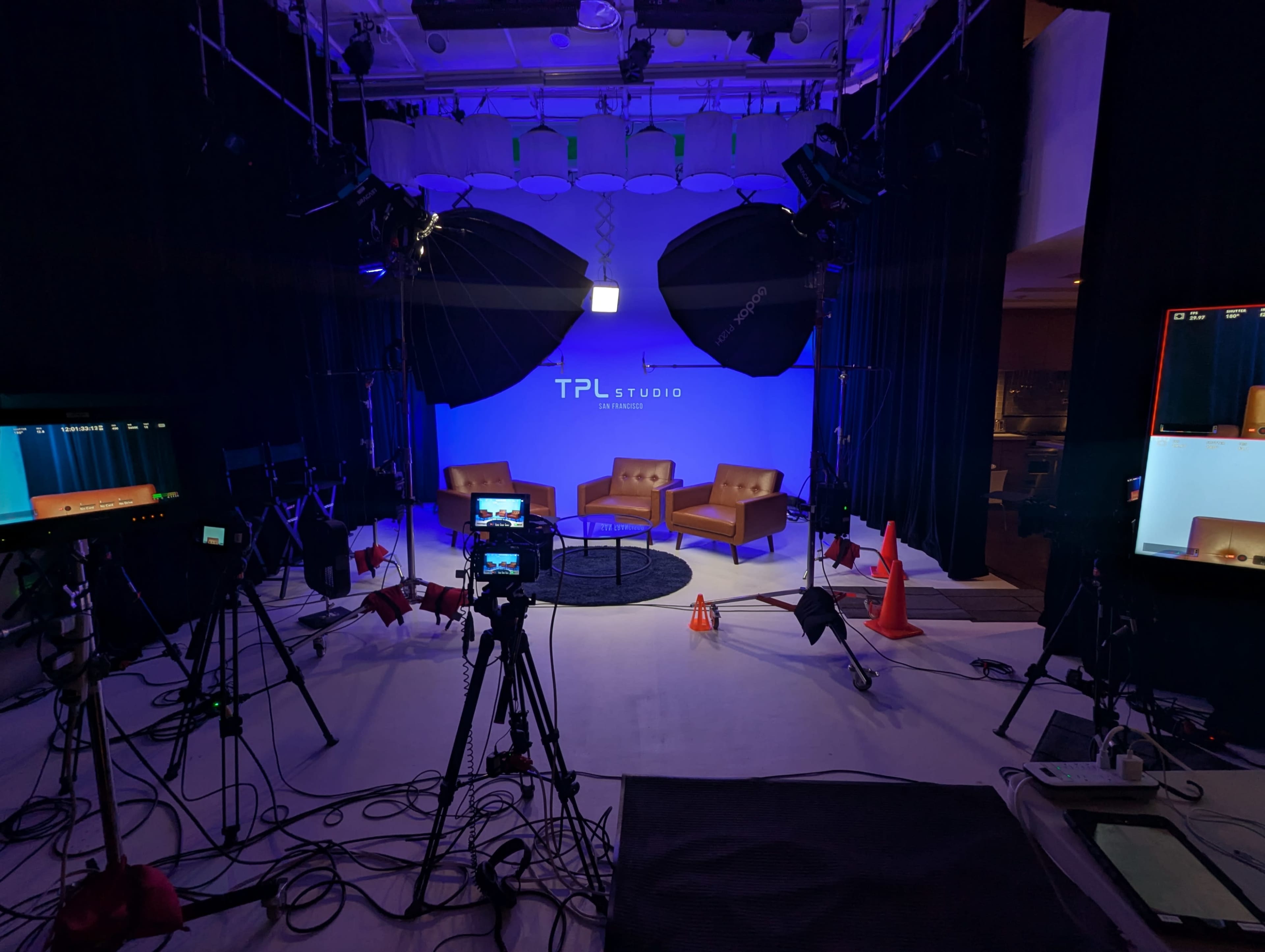 The image shows a film set with two brown chairs on a circular rug, surrounded by cameras, lights, and equipment against a blue backdrop with the text "TPL Studio."
