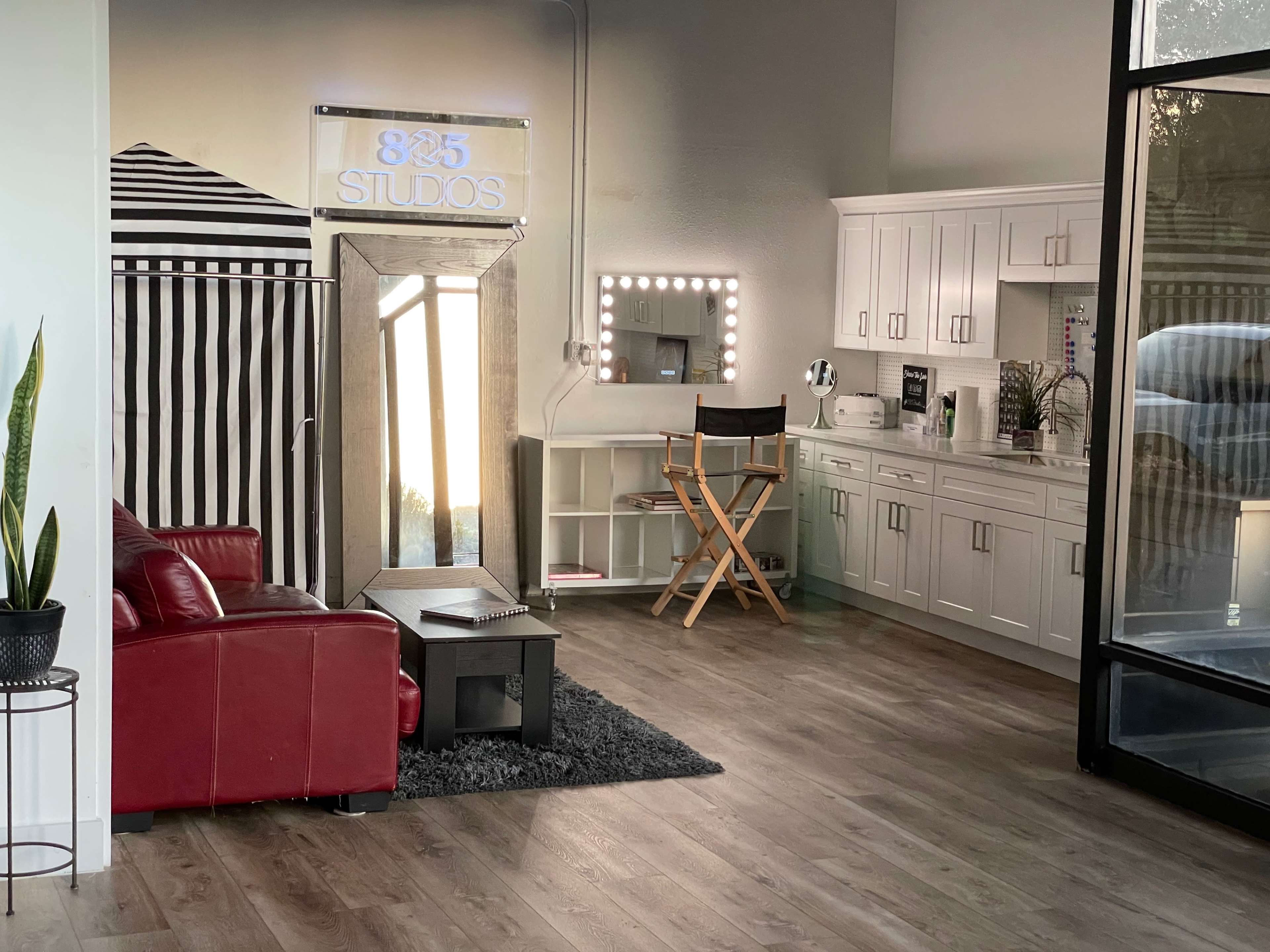 A modern studio space featuring a red sofa, a mirrored vanity with lights, and a white cabinetry setup against a backdrop with a striped pattern.