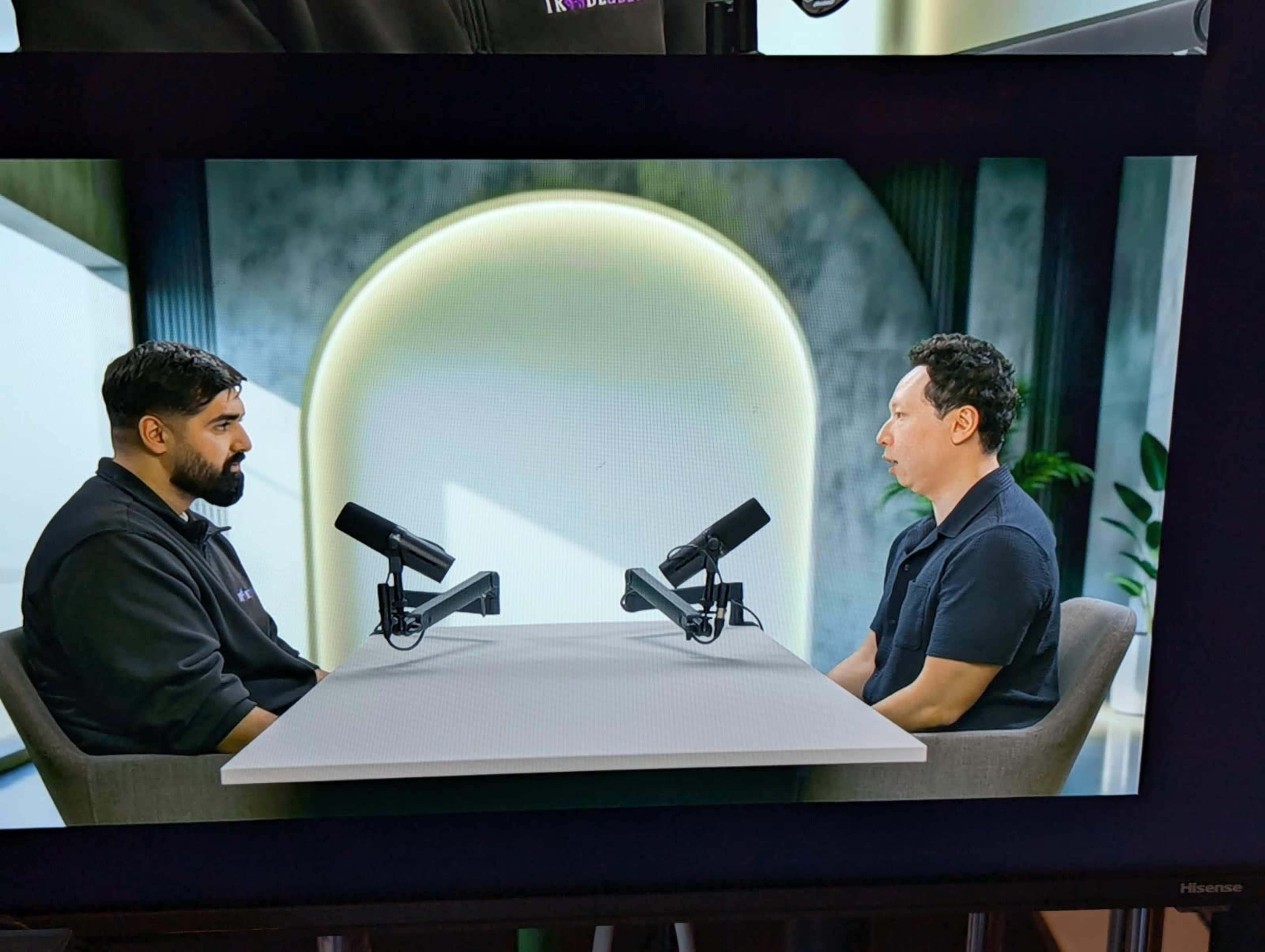 Two men are seated at a table with microphones in front of them, engaged in a conversation against a modern, minimalist background.