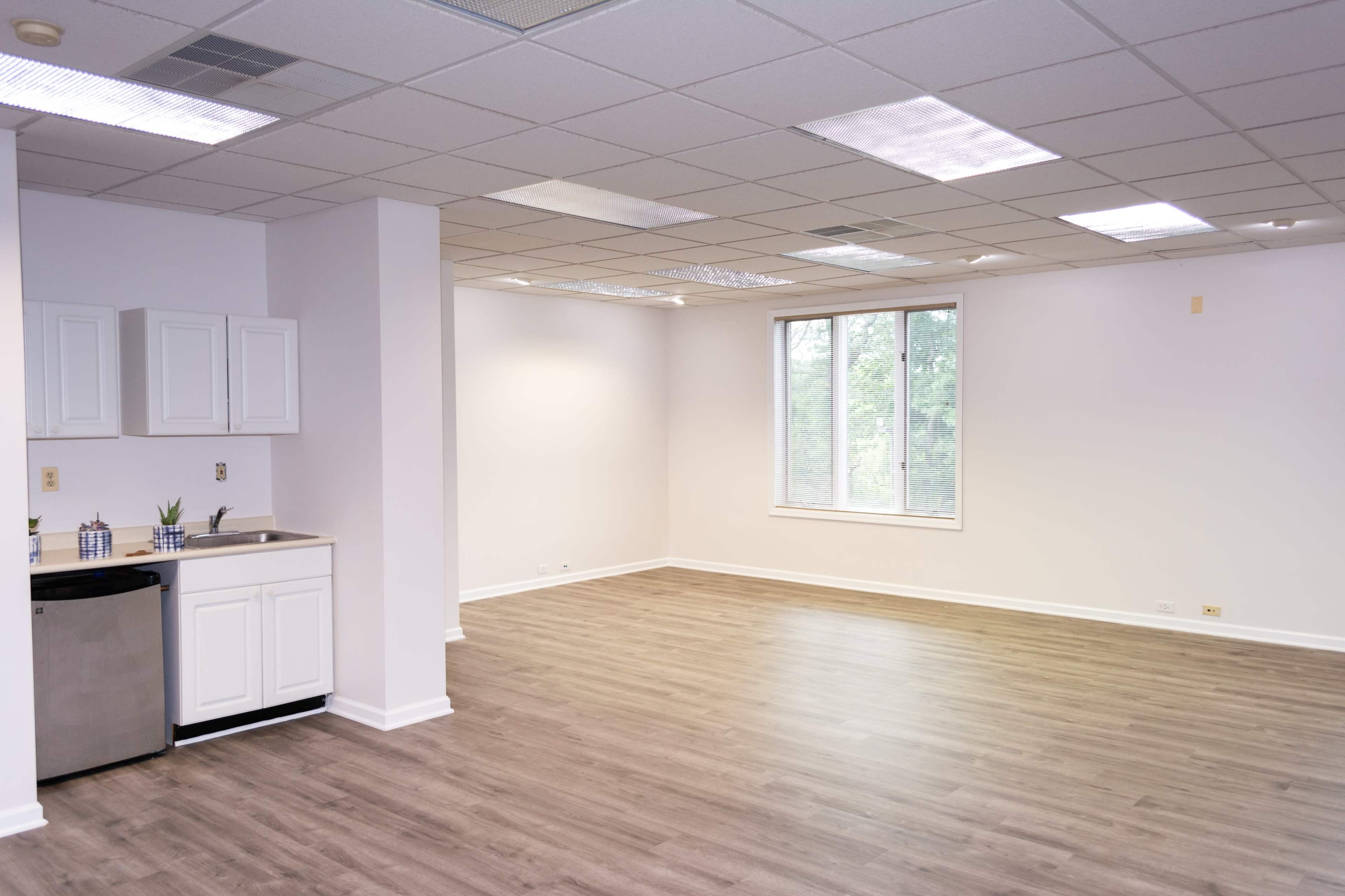 An empty, well-lit room with wooden flooring, a kitchen area featuring a small refrigerator and white cabinets, and large windows providing natural light.