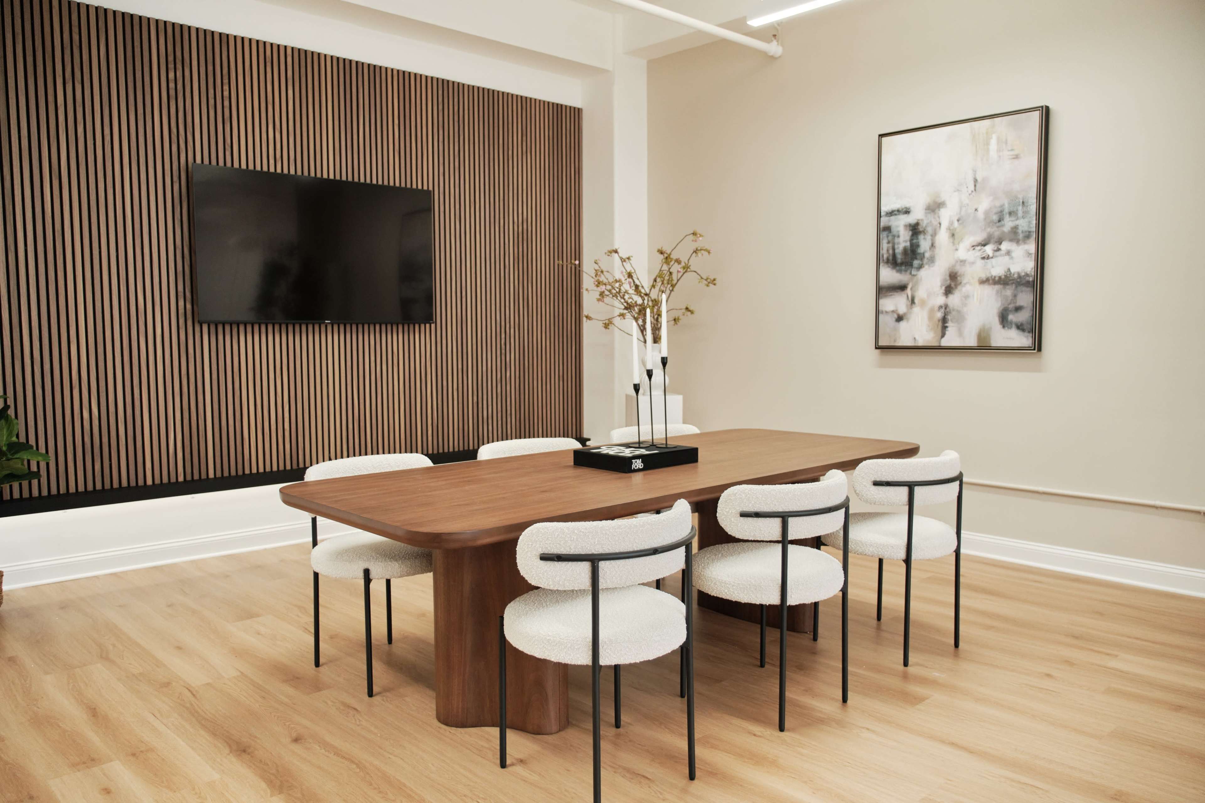 A modern conference room features a wooden table surrounded by upholstered chairs, a wall-mounted television, and artwork.