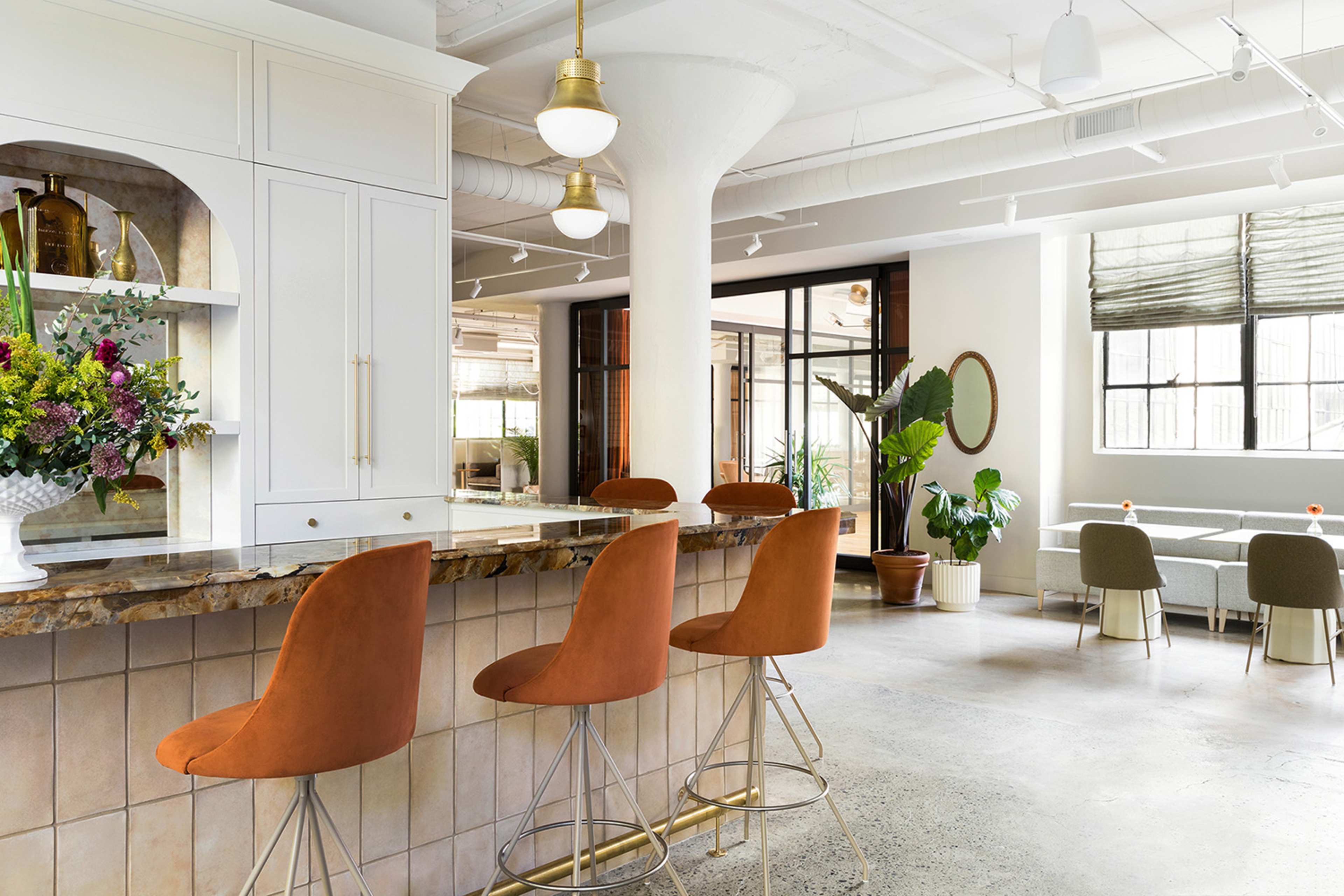 A modern kitchen area with a bar featuring four orange stools, a countertop with a floral arrangement, and a dining space with green chairs in the background.