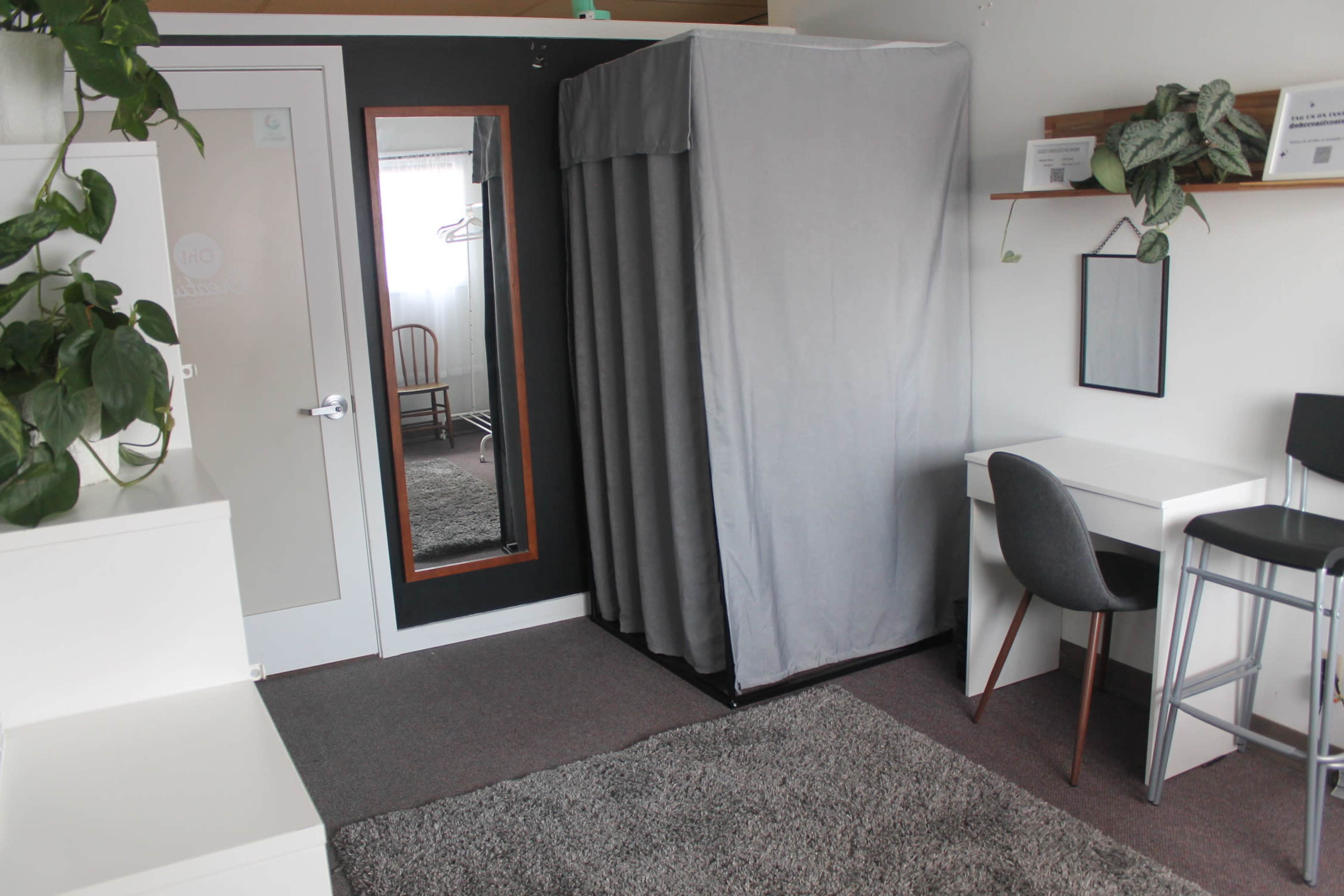 A small room featuring a gray privacy curtain, a mirror, a desk with a chair, and a wooden chair against the wall, along with potted plants and a gray rug on the floor.