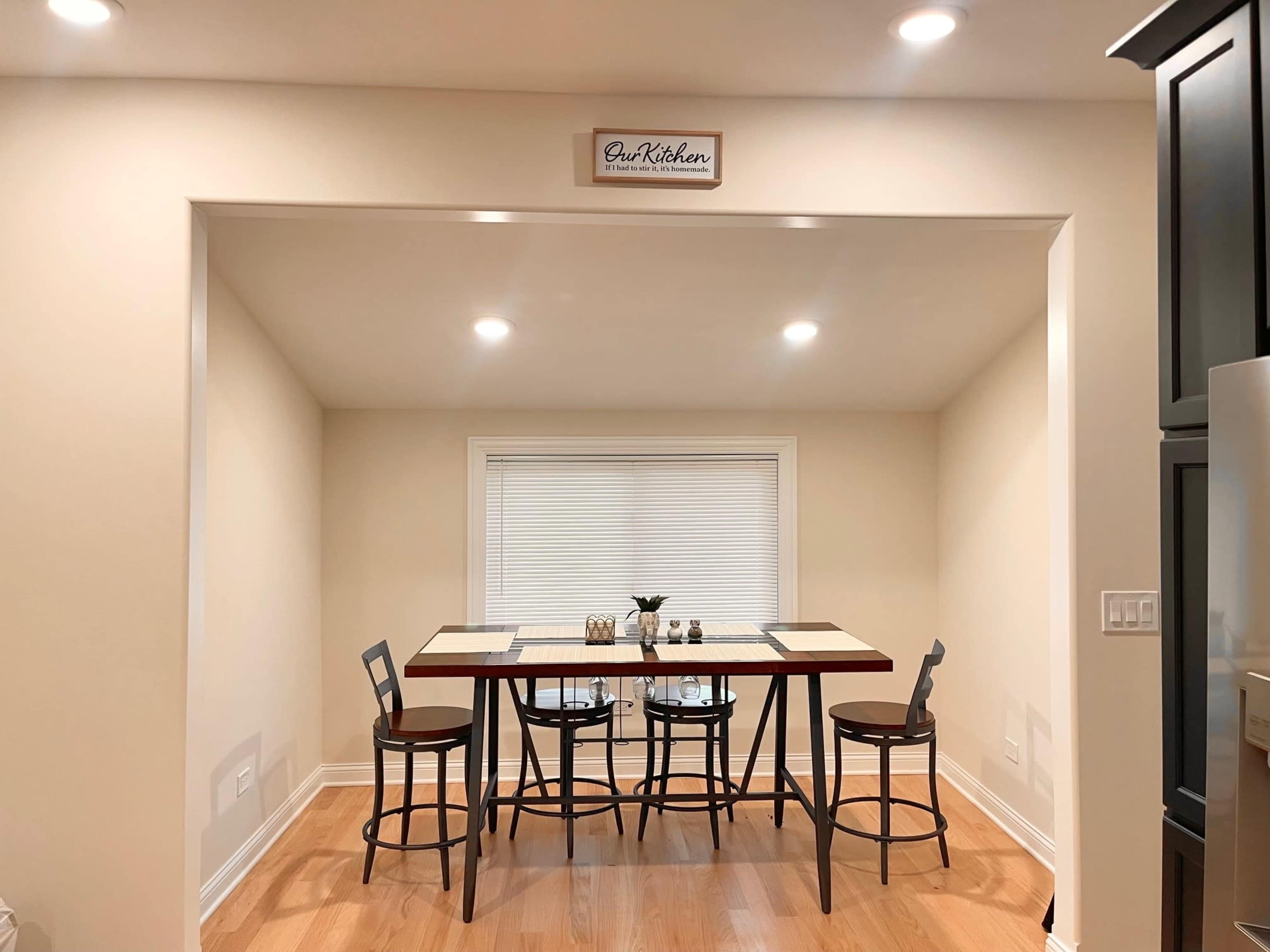 A small dining area features a table with four chairs and a window above, illuminated by overhead lights.