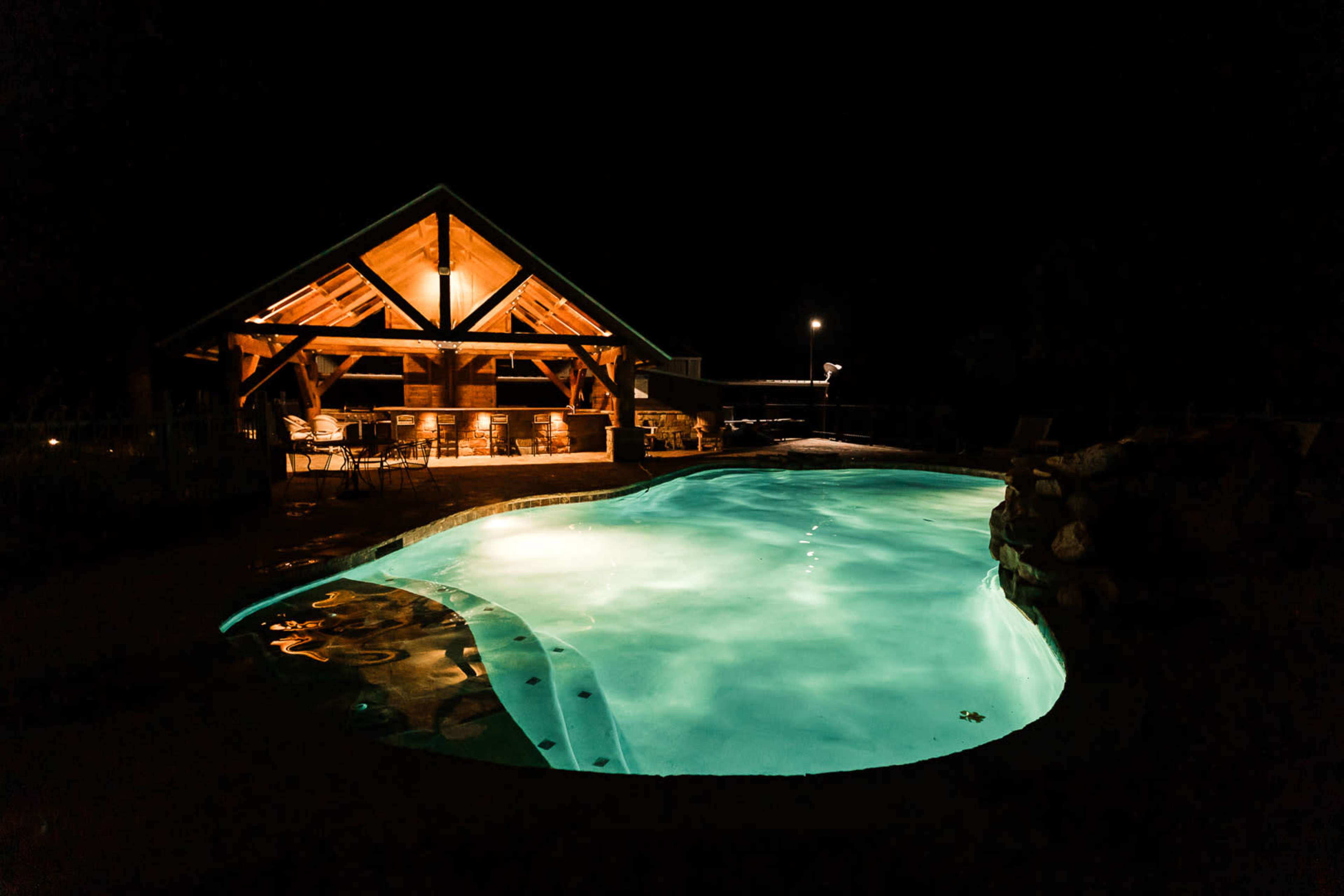 A brightly lit swimming pool reflects the light from a nearby wooden pavilion at night.