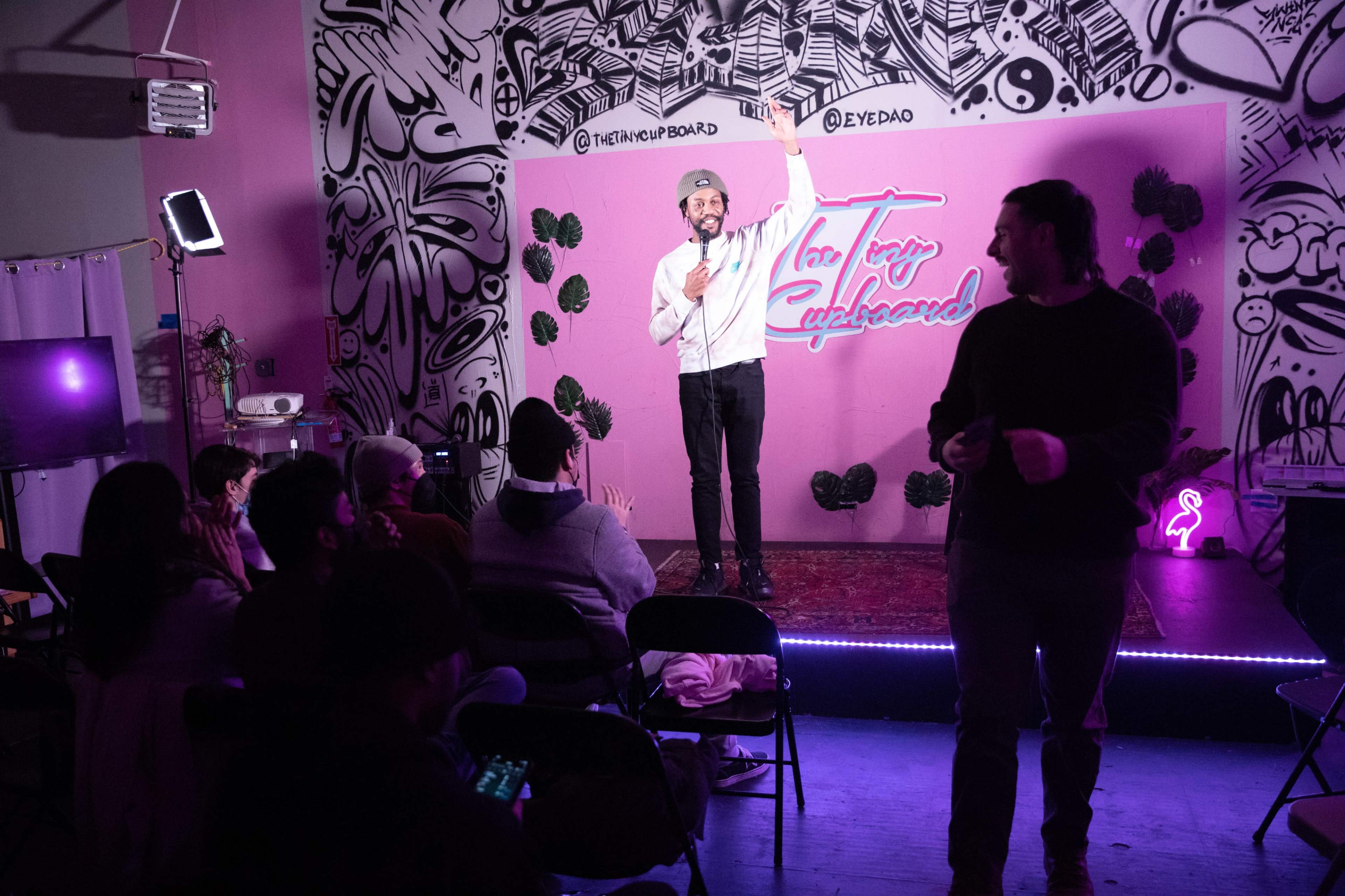 A comedian stands on stage with a microphone, addressing an audience in a colorful room decorated with graffiti.