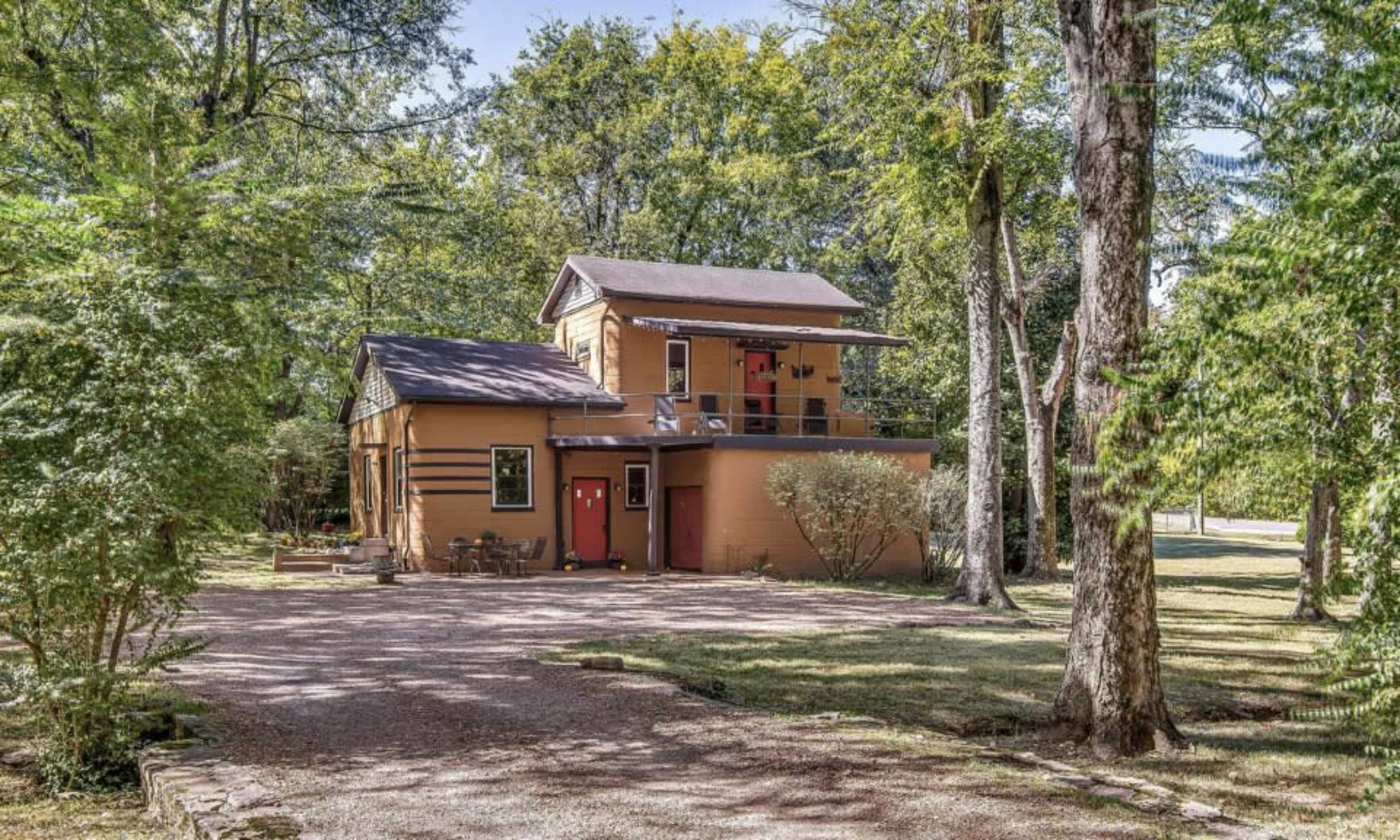 A two-story house with a balcony and red doors is surrounded by trees and gravel pathways.