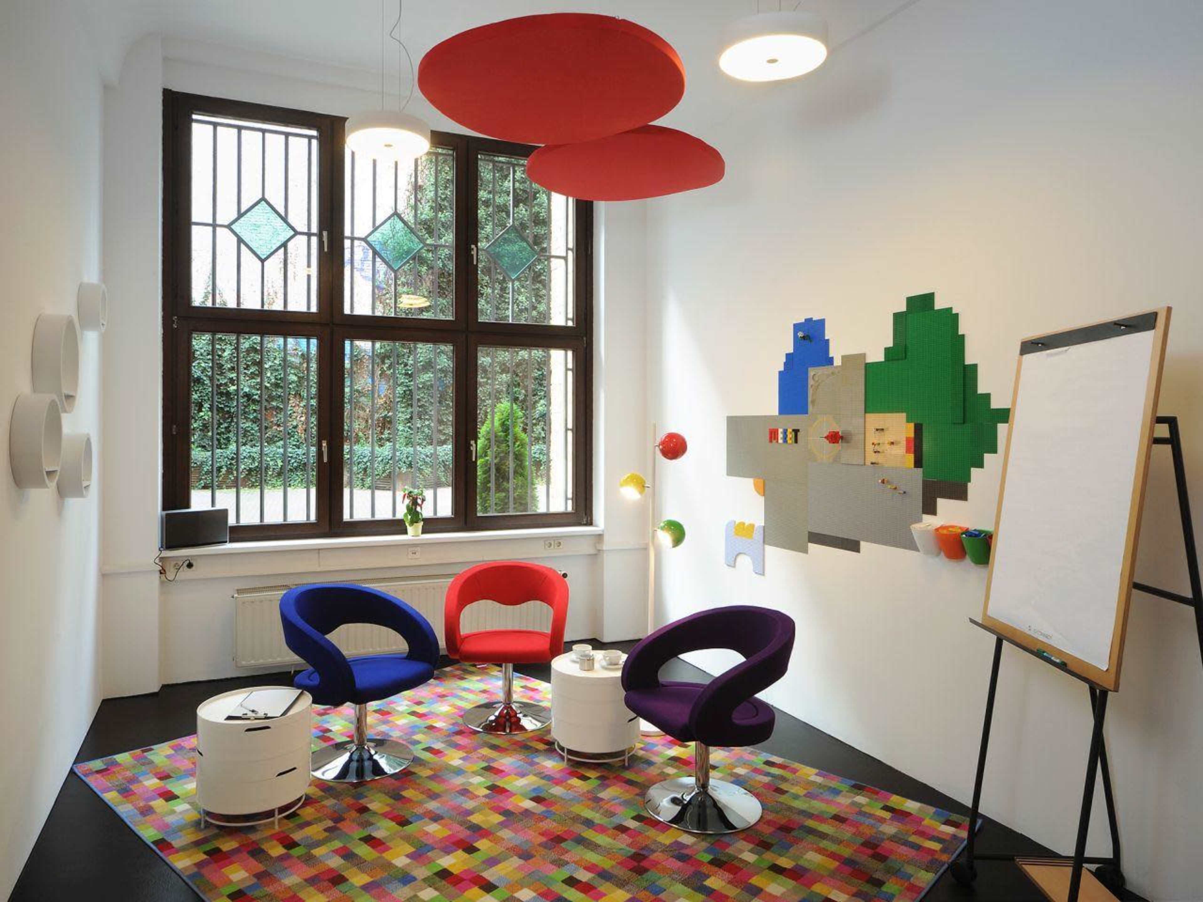 A modern meeting room featuring colorful chairs, a patterned rug, a large window with greenery outside, and a wall with abstract decorations.