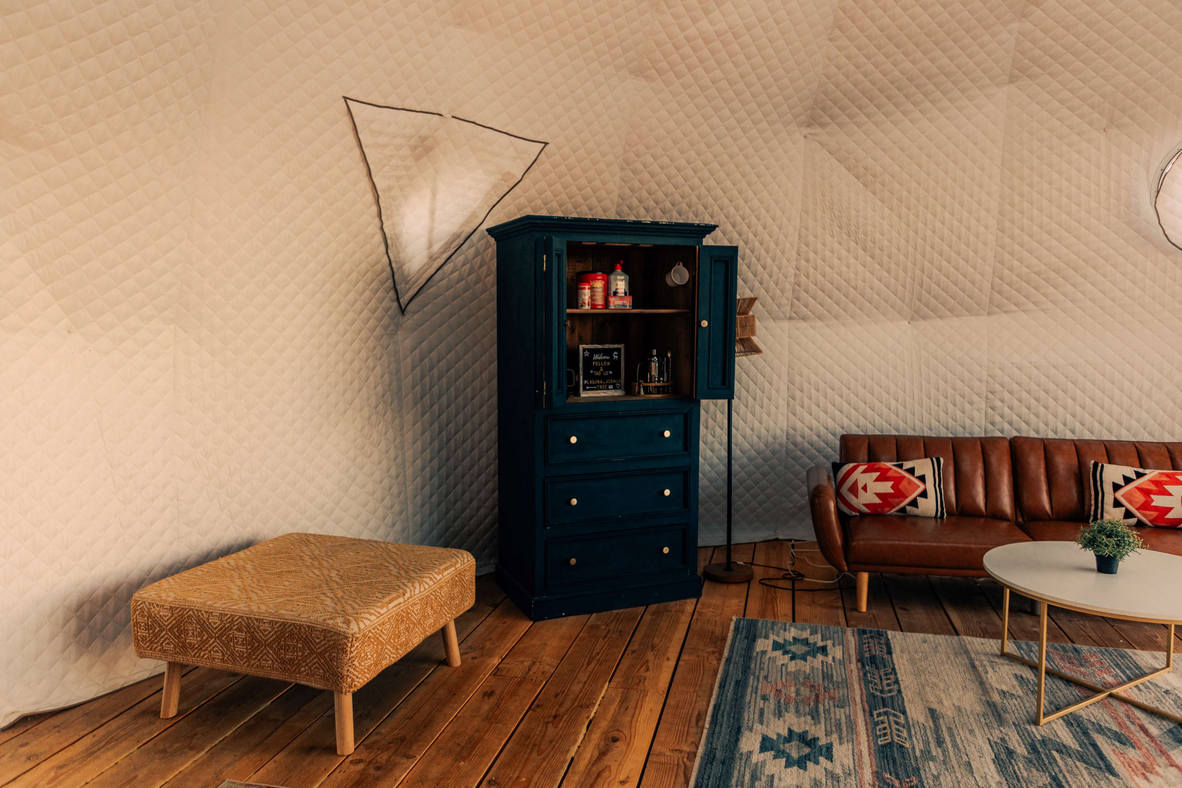 The image shows a cozy interior featuring a blue wooden cabinet, a brown leather sofa with decorative cushions, a round coffee table with a small plant, and a patterned rug on a wooden floor.