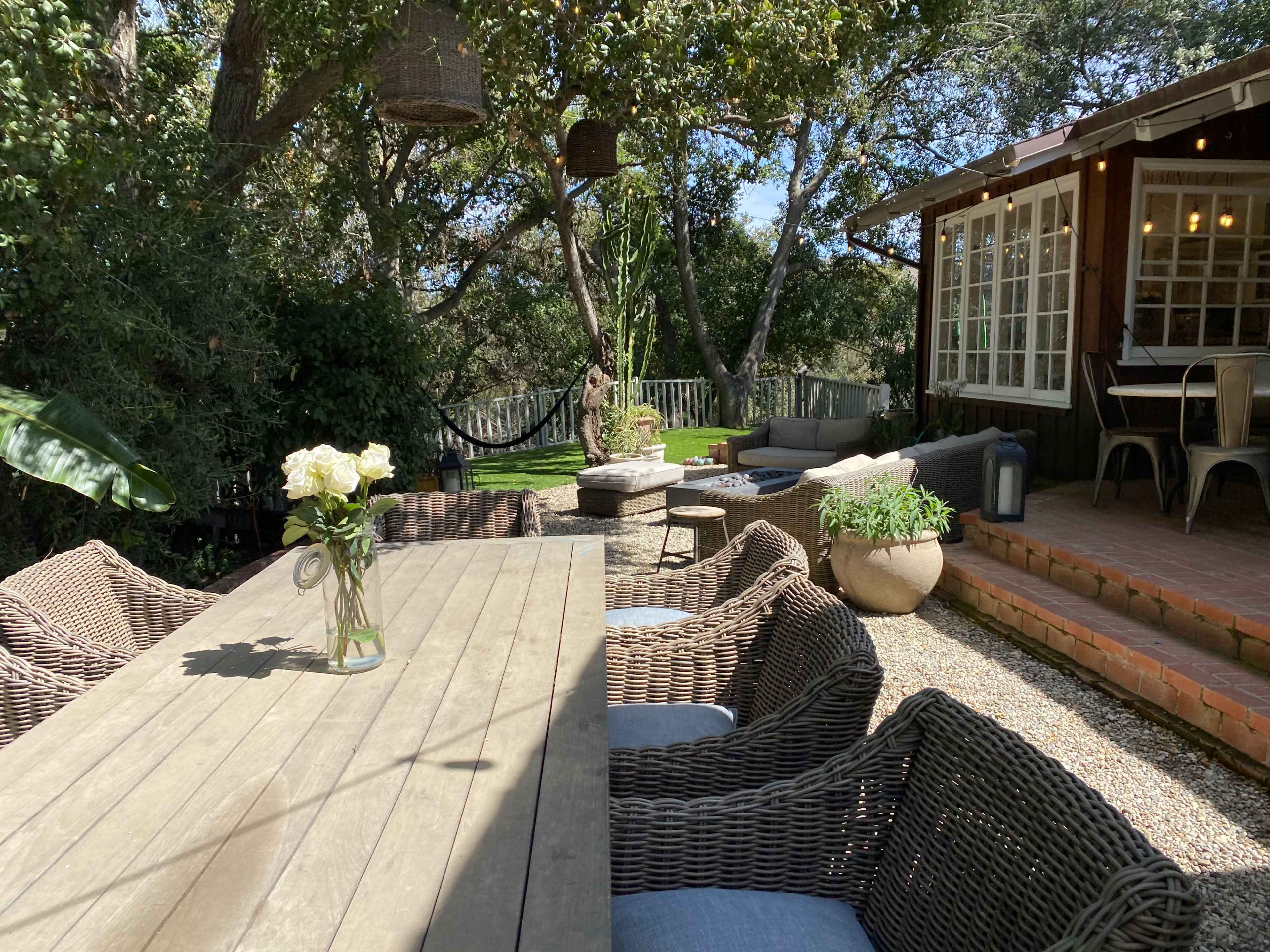 A wooden dining table with wicker chairs is set on a gravel patio beside a house surrounded by trees and greenery.