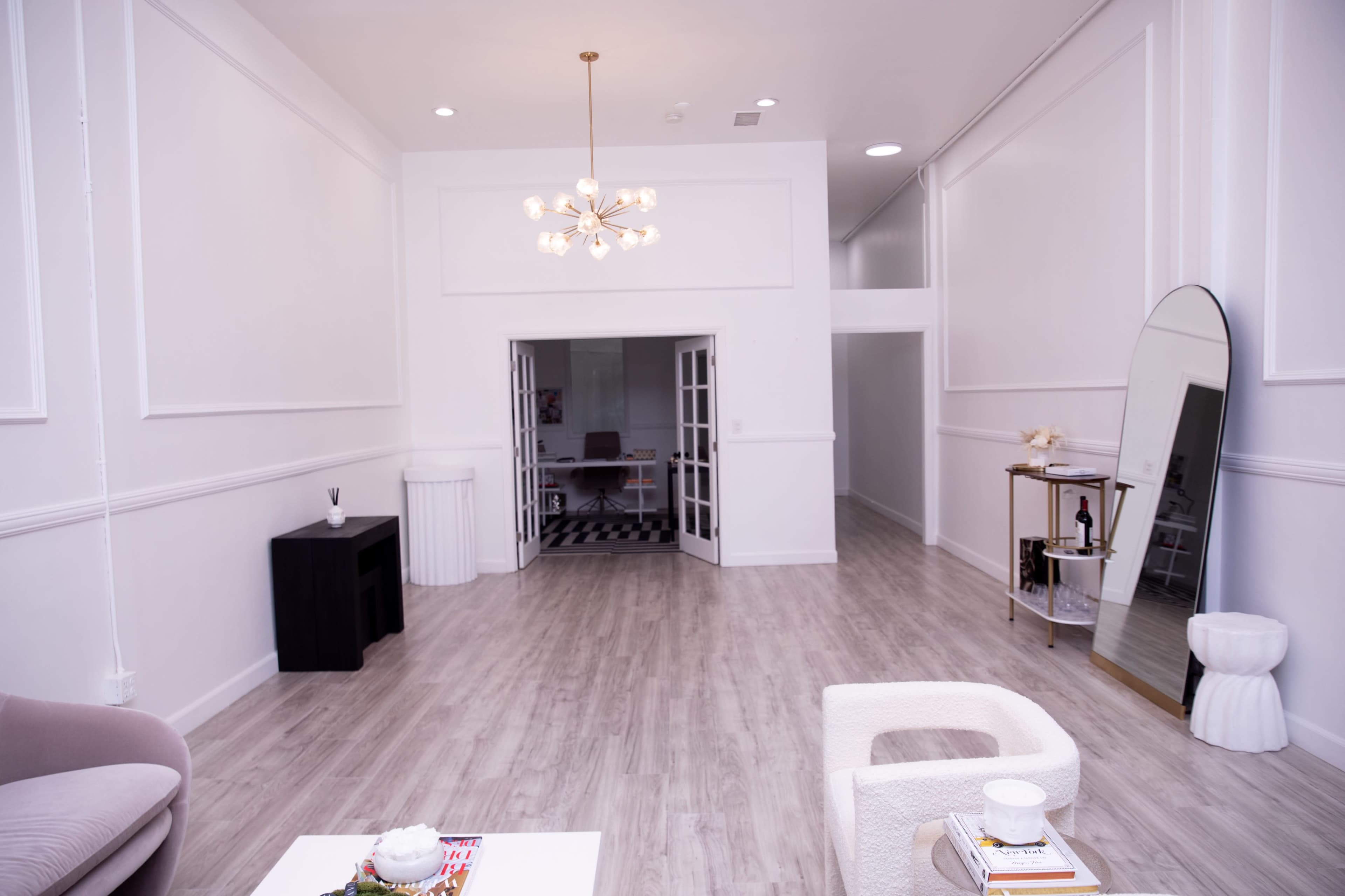 A spacious, empty room features light wood flooring, white walls, and modern furniture, including a black side table and a large mirror, with doors leading to another room in the background.