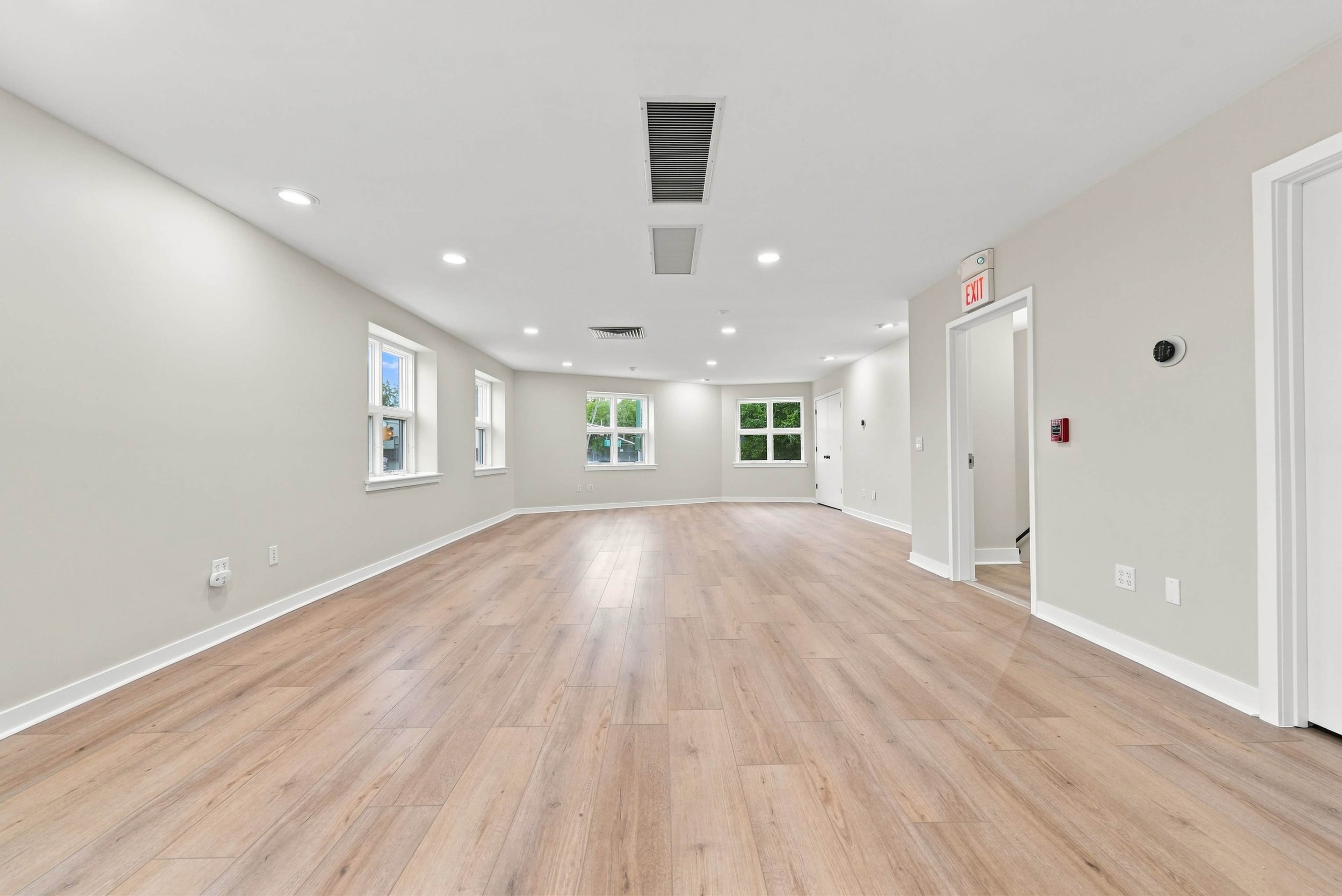 The image shows a spacious, empty room with light wood flooring and several windows allowing natural light to enter.