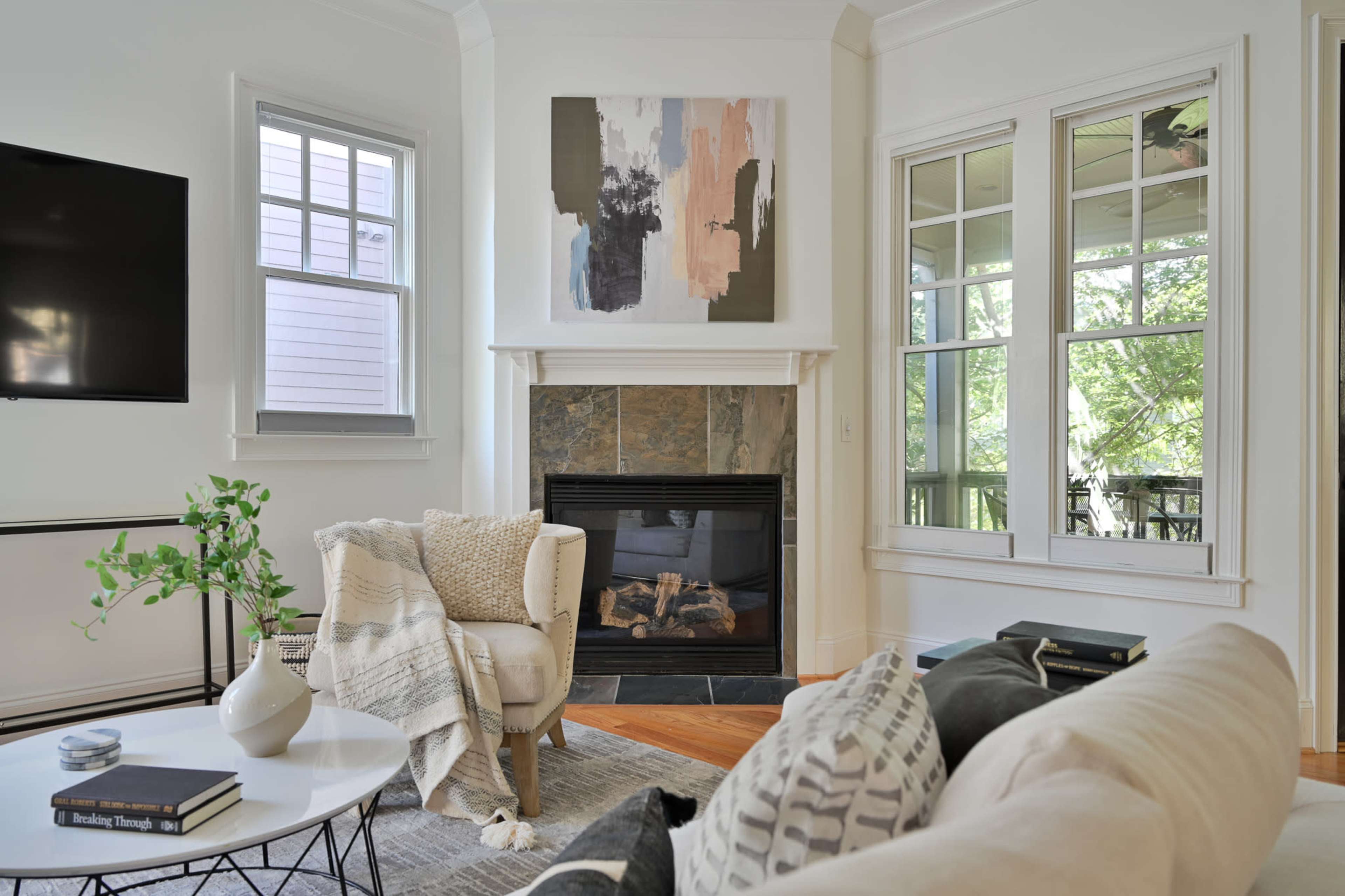 A bright living room featuring a fireplace, a large abstract painting on the wall, a coffee table with books, and large windows that overlook a green outdoor space.