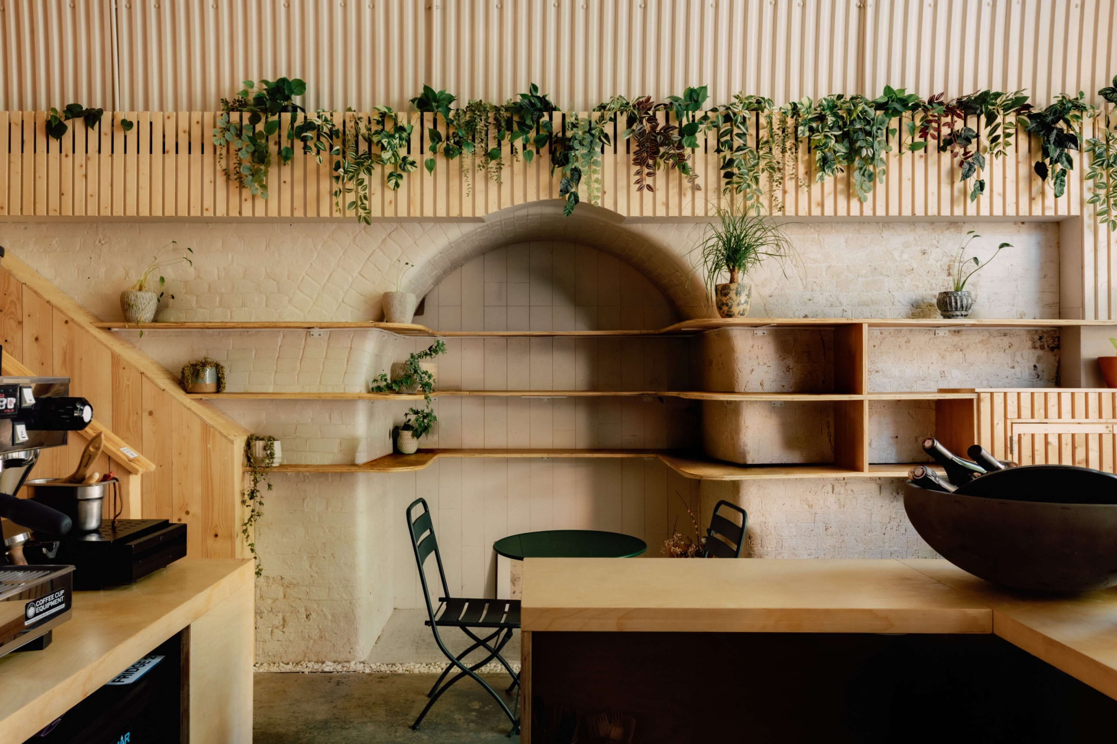 The image shows a minimalist café interior featuring wooden shelves adorned with plants, a small green table with two chairs, and a coffee machine on the counter.
