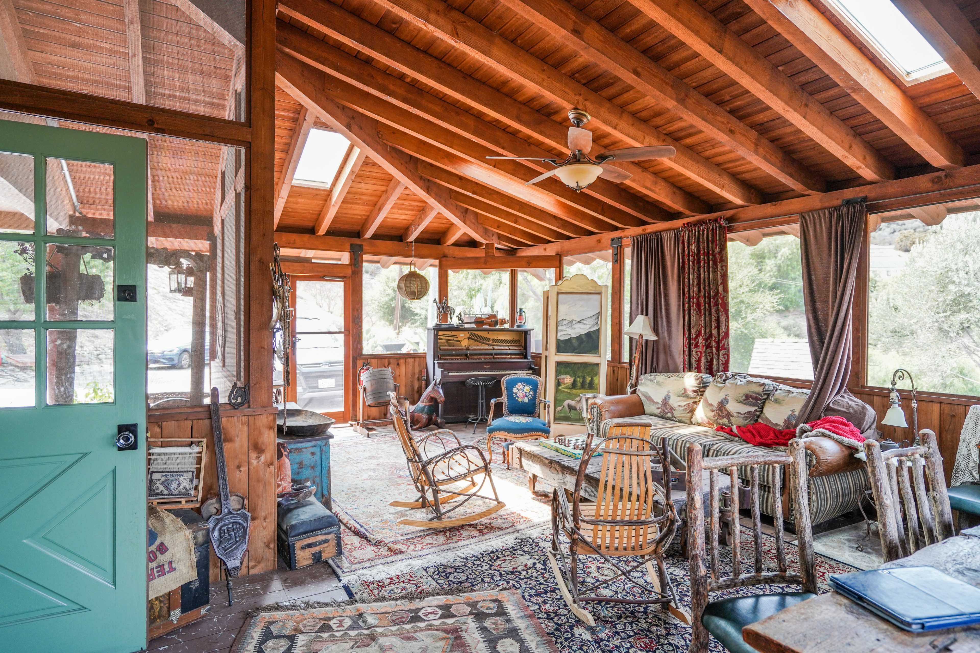 The photo shows a rustic living room with wooden beams, large windows, and a combination of vintage furniture, including a sofa, rocking chairs, and a decorative rug.