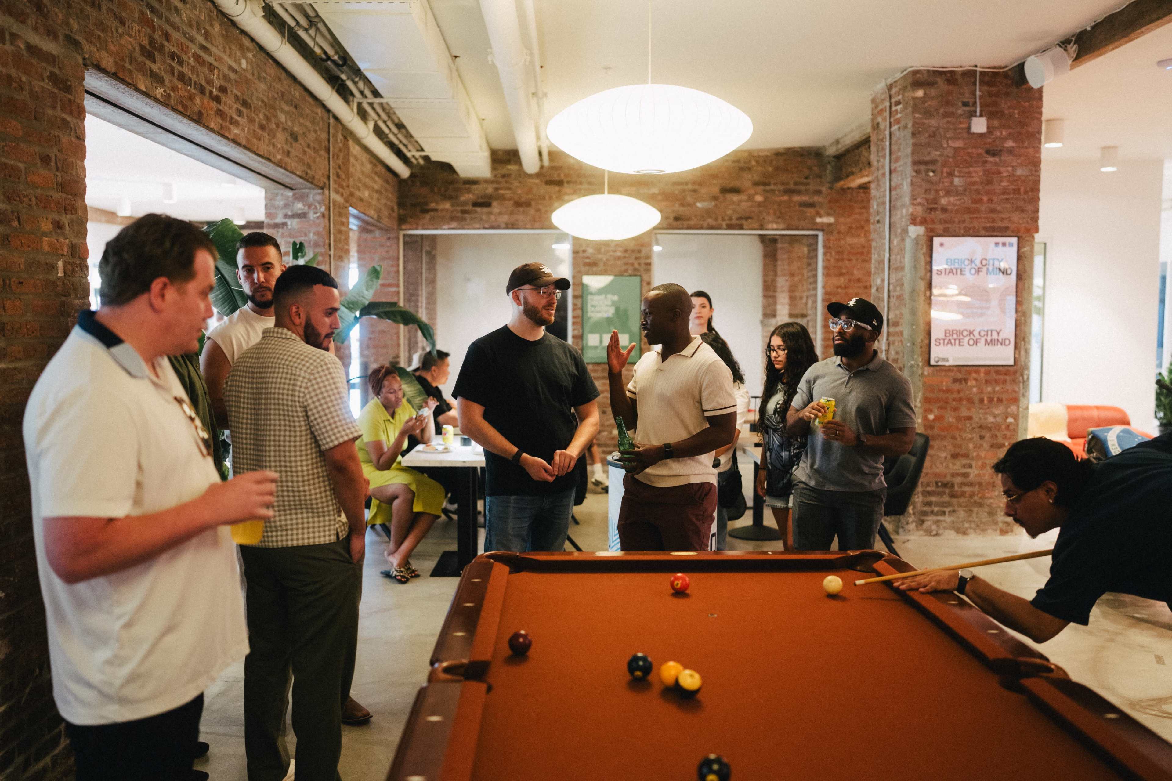 A group of people gathers around a pool table in a casual indoor setting with exposed brick walls.