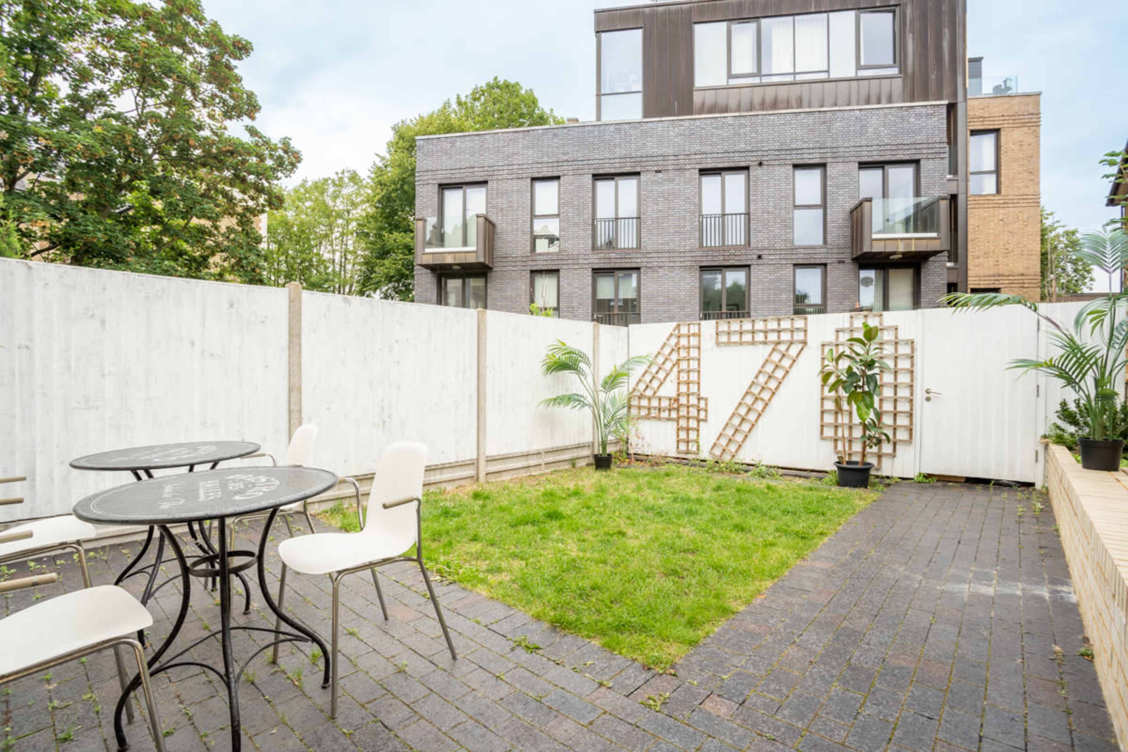 A small outdoor area features a grassy space surrounded by wooden fencing, with a table and chairs set on a stone patio beside a modern building.