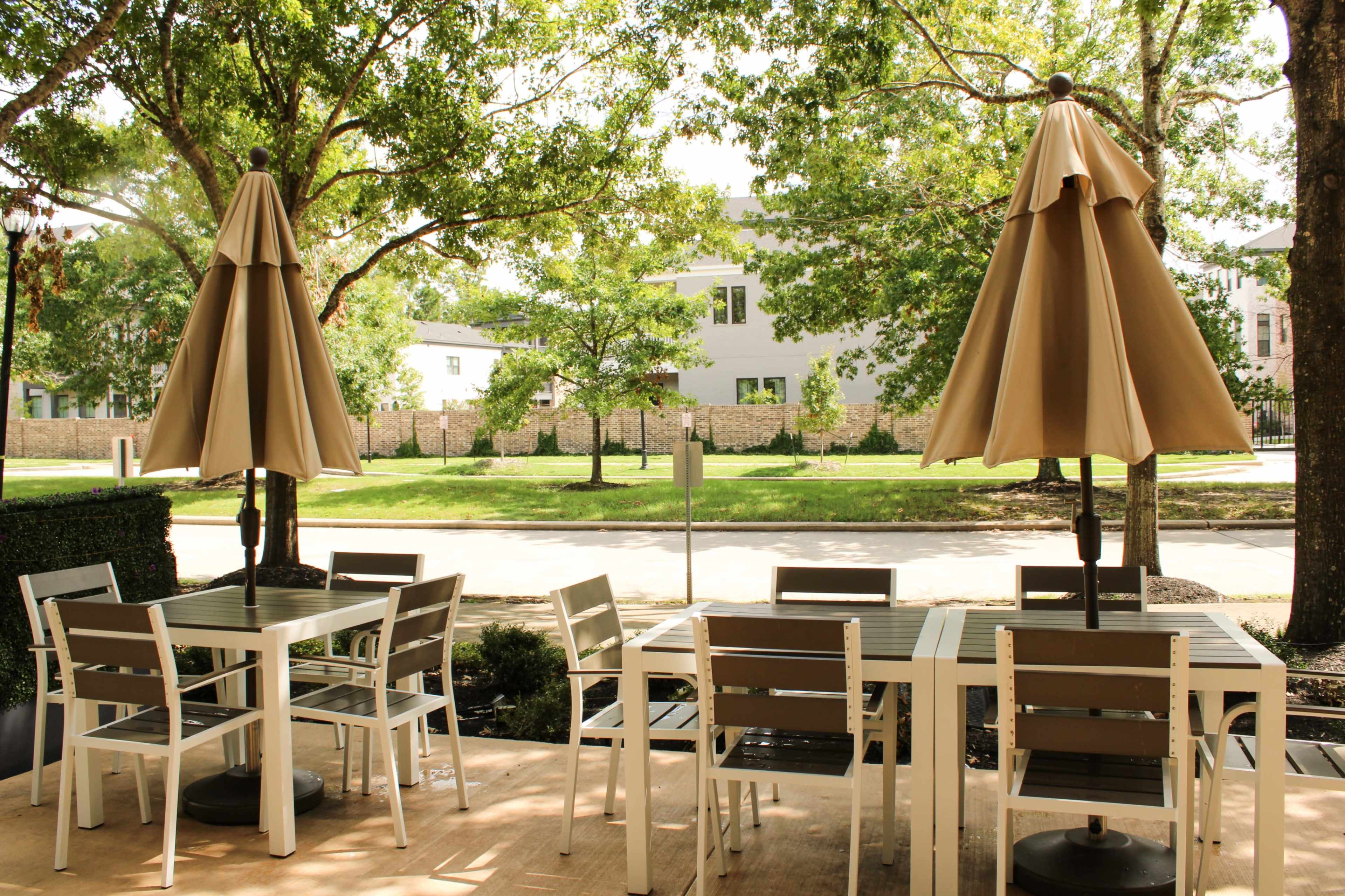 An outdoor seating area with white tables and brown chairs is shaded by large umbrellas, overlooking a tree-lined street.