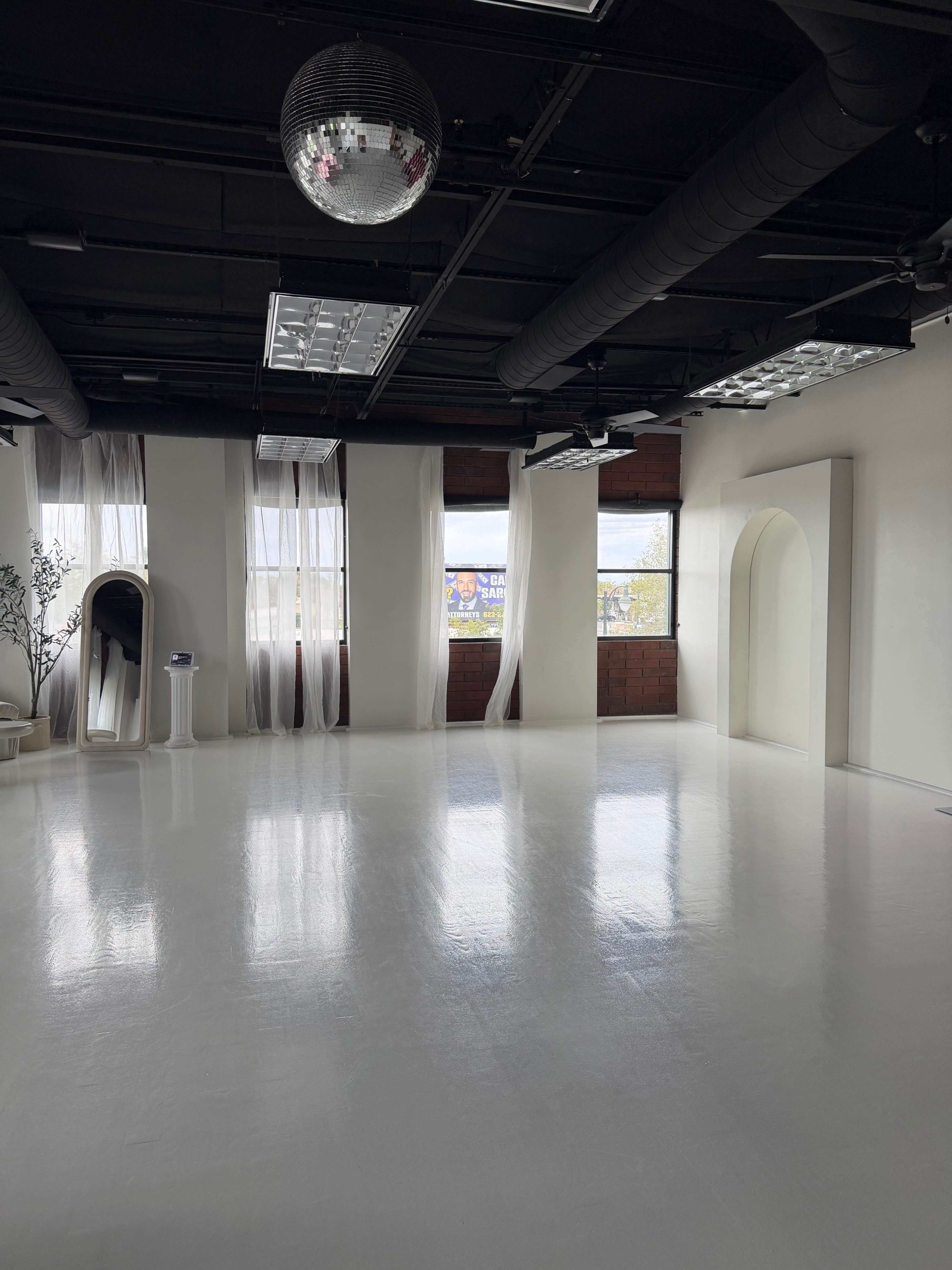 The image shows a spacious, well-lit room with white floors, large windows, and a disco ball hanging from the ceiling.