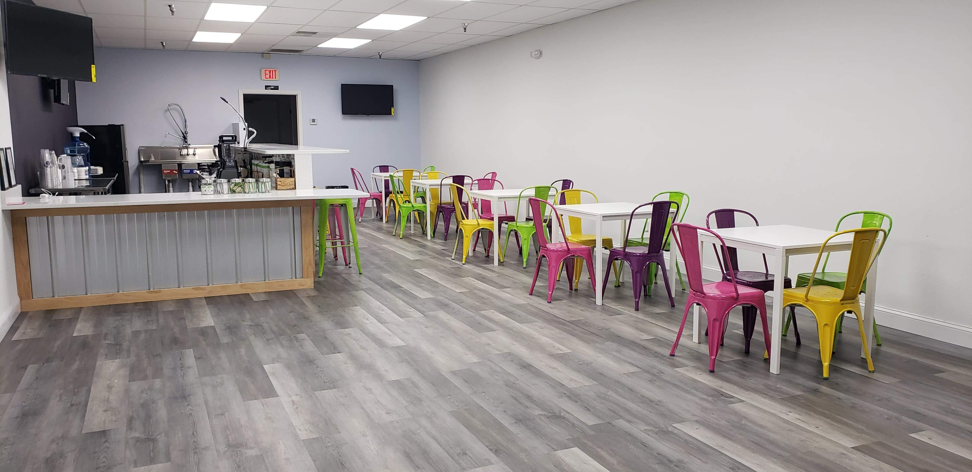 A brightly colored café features white tables surrounded by multi-colored metal chairs on a gray floor.