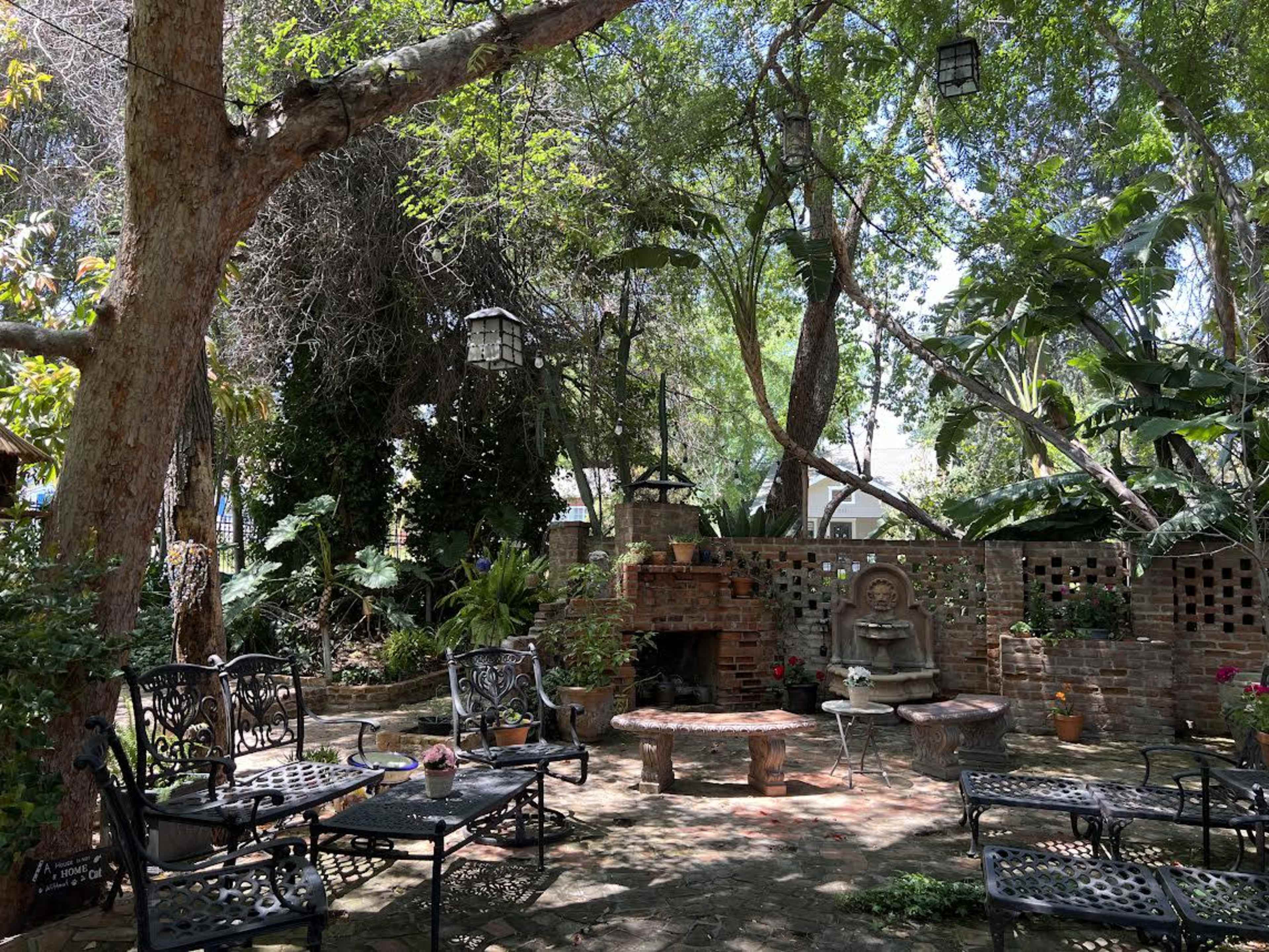 The scene features a landscaped garden with stone pathways, a central fountain, lush greenery, and metal chairs arranged around a table.
