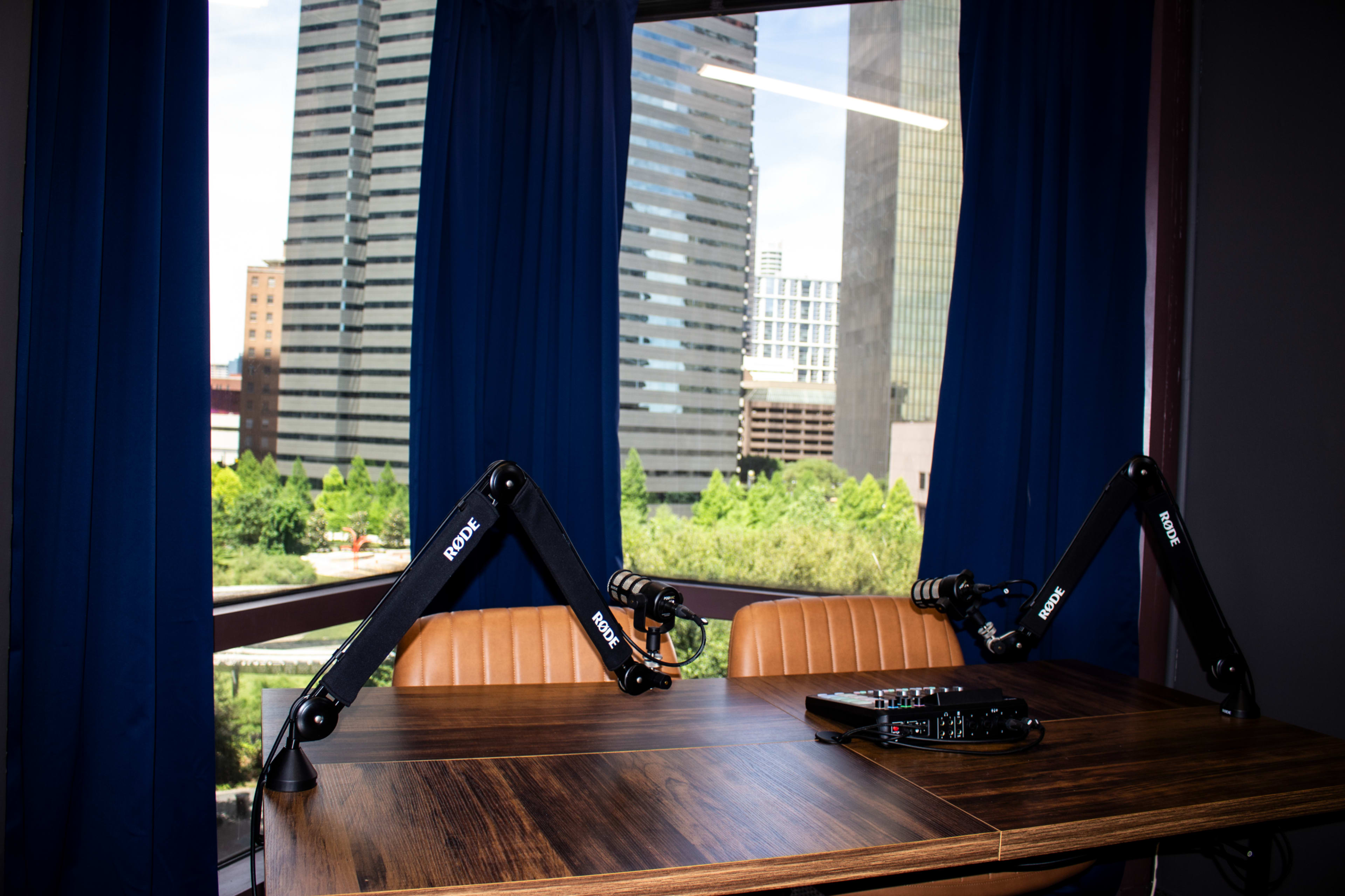 The image shows a recording studio setup with two microphone arms extended over a wooden table, facing a large window that overlooks city buildings.