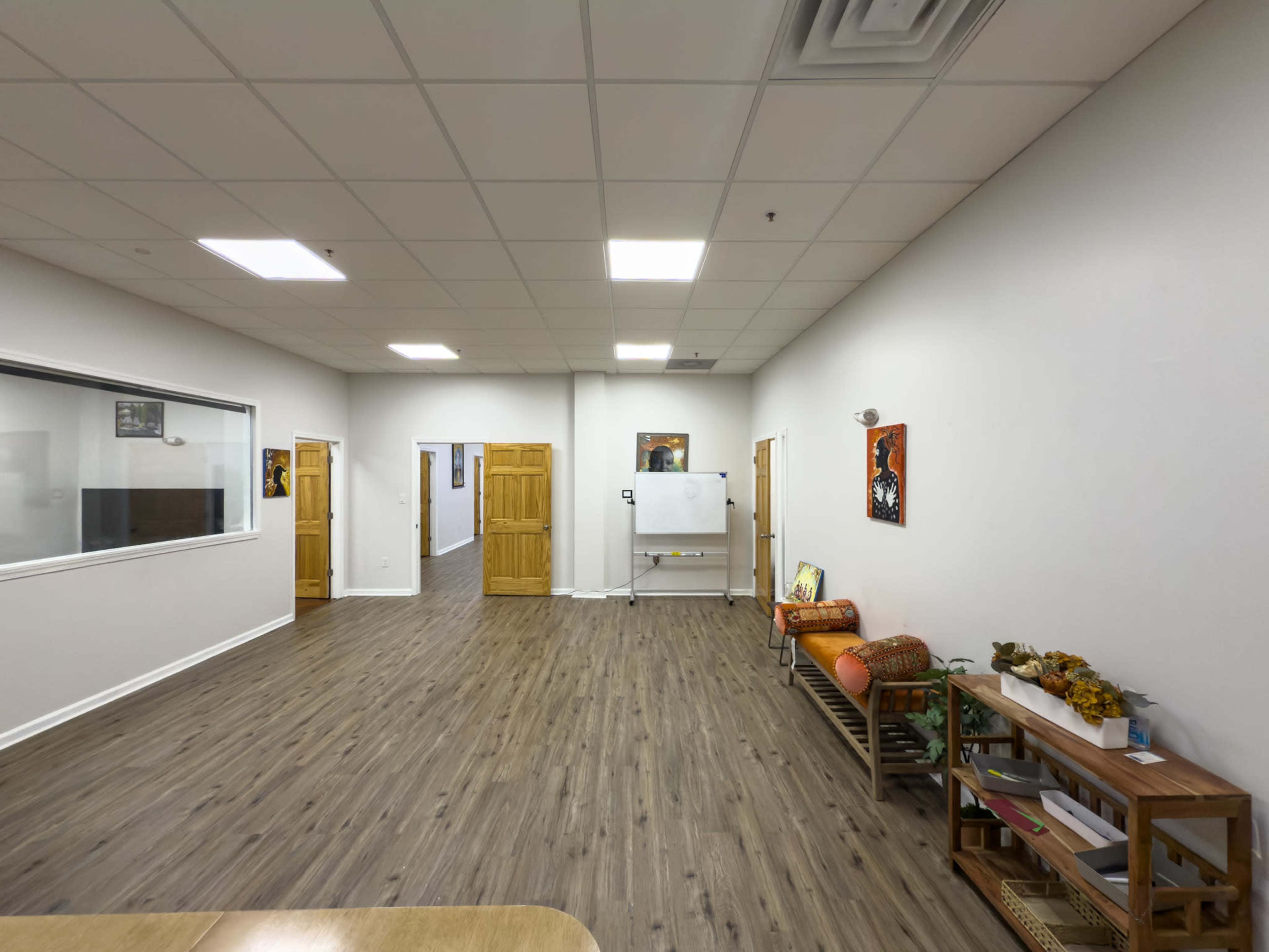 The image shows a spacious, well-lit room with wooden flooring, featuring a whiteboard, a small seating area, and art displayed on the walls.