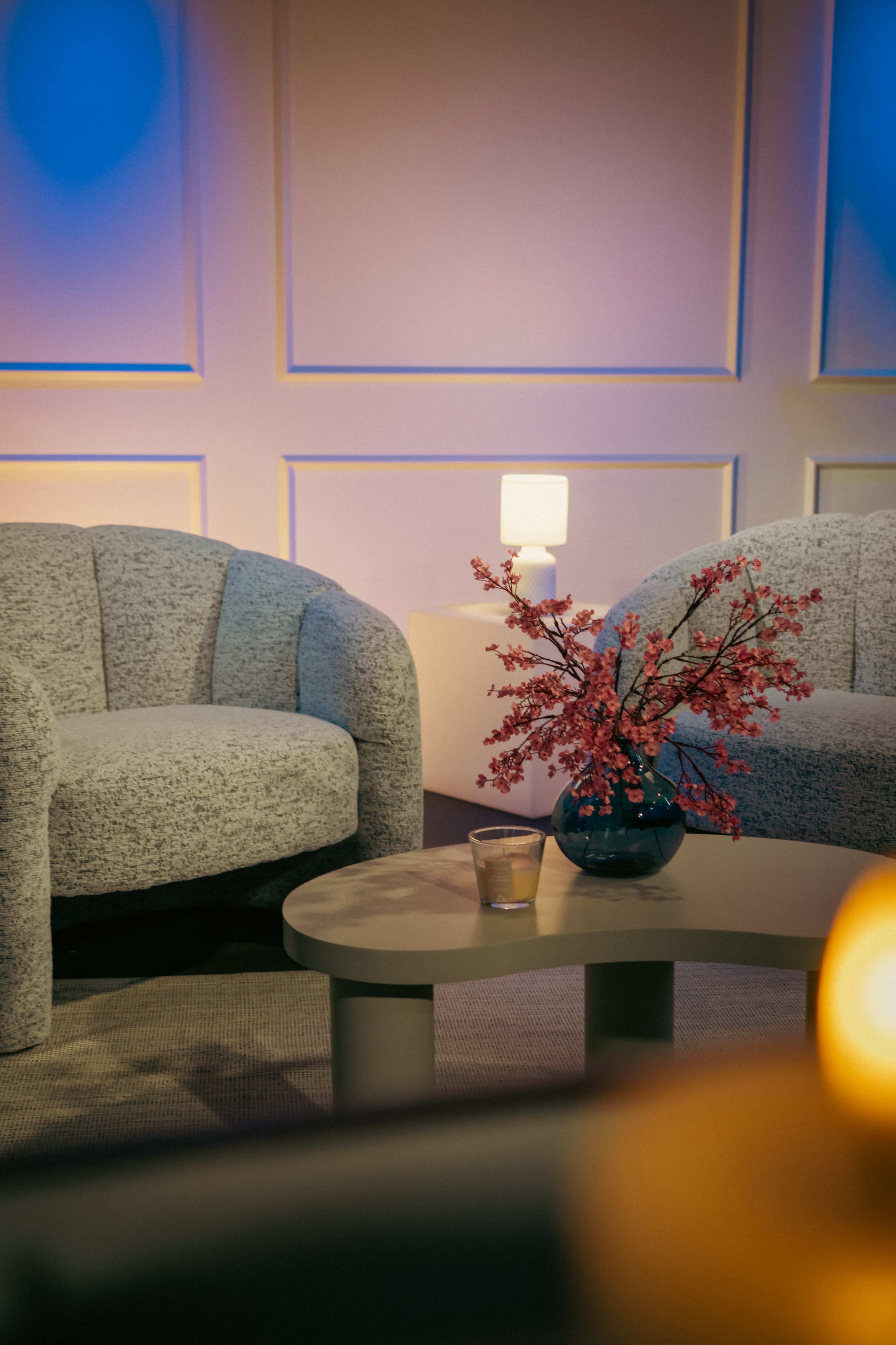 The image features two upholstered chairs and a round coffee table with a small vase of pink flowers and a glass on it, set against a softly lit background.