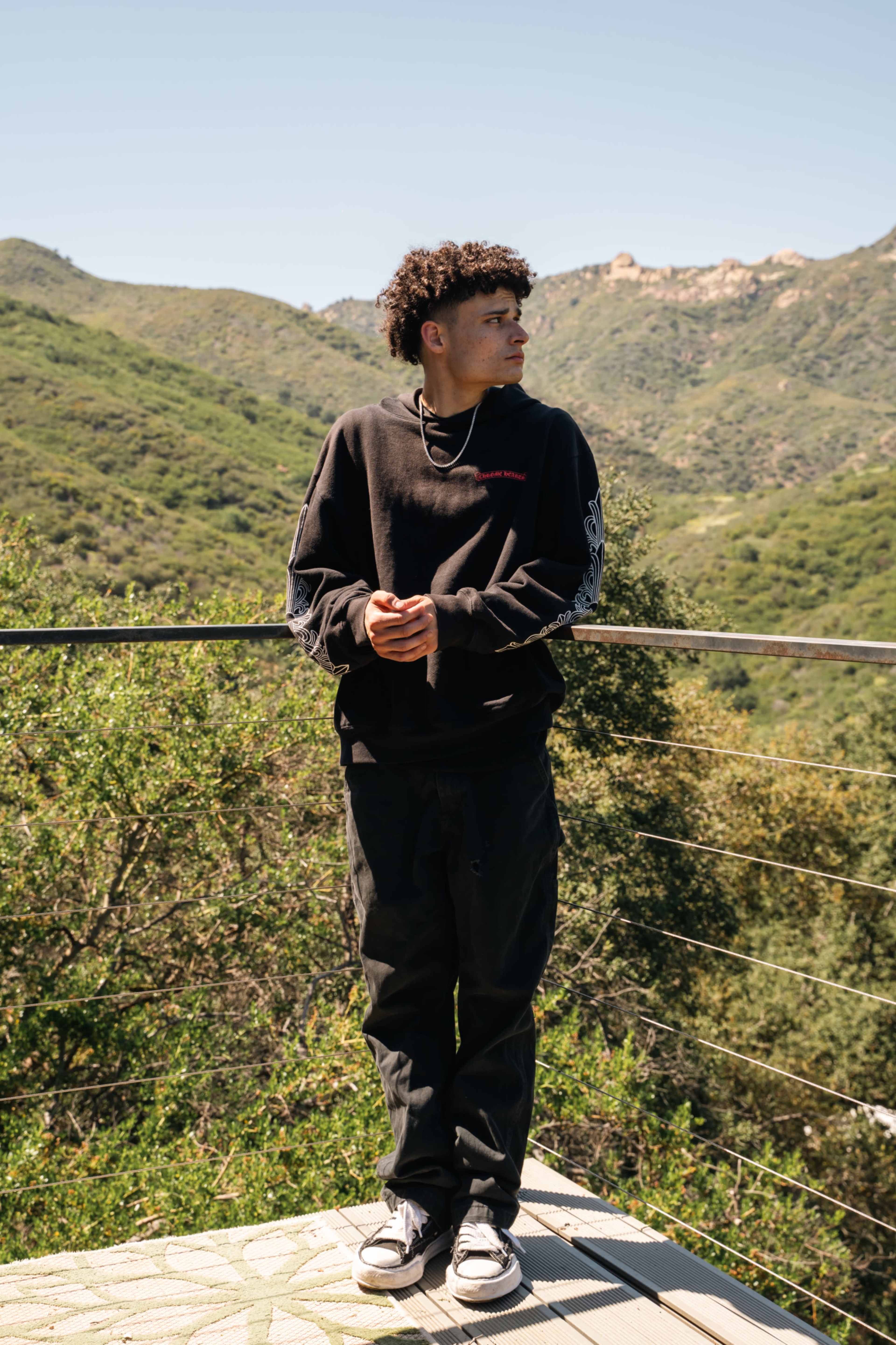 A young person stands on a terrace overlooking a green mountainous landscape under a clear blue sky.