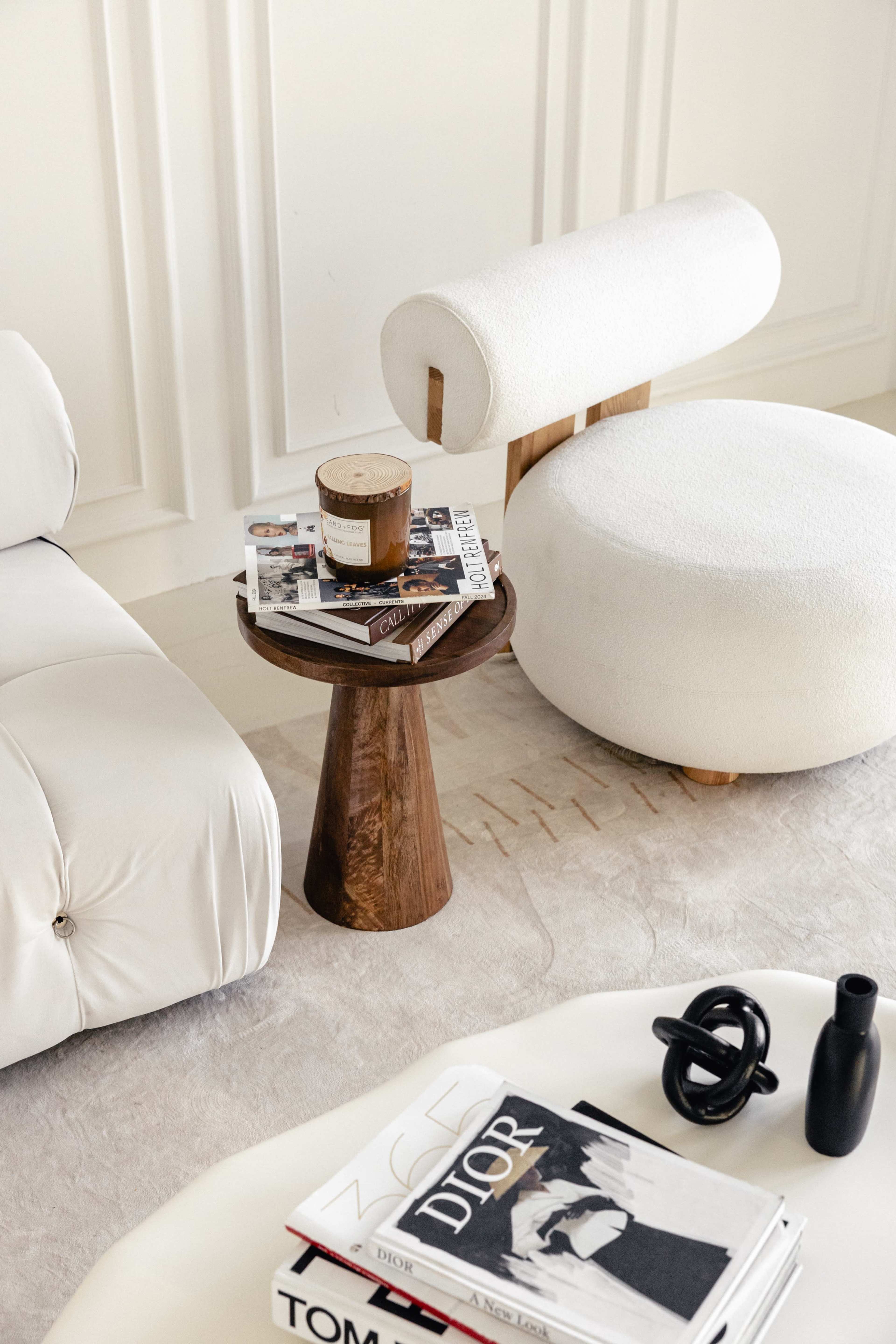 The image features a modern living room setting with a cream-colored sofa, a circular stool, a wooden side table holding magazines and a candle, and a decorative black object on a coffee table.