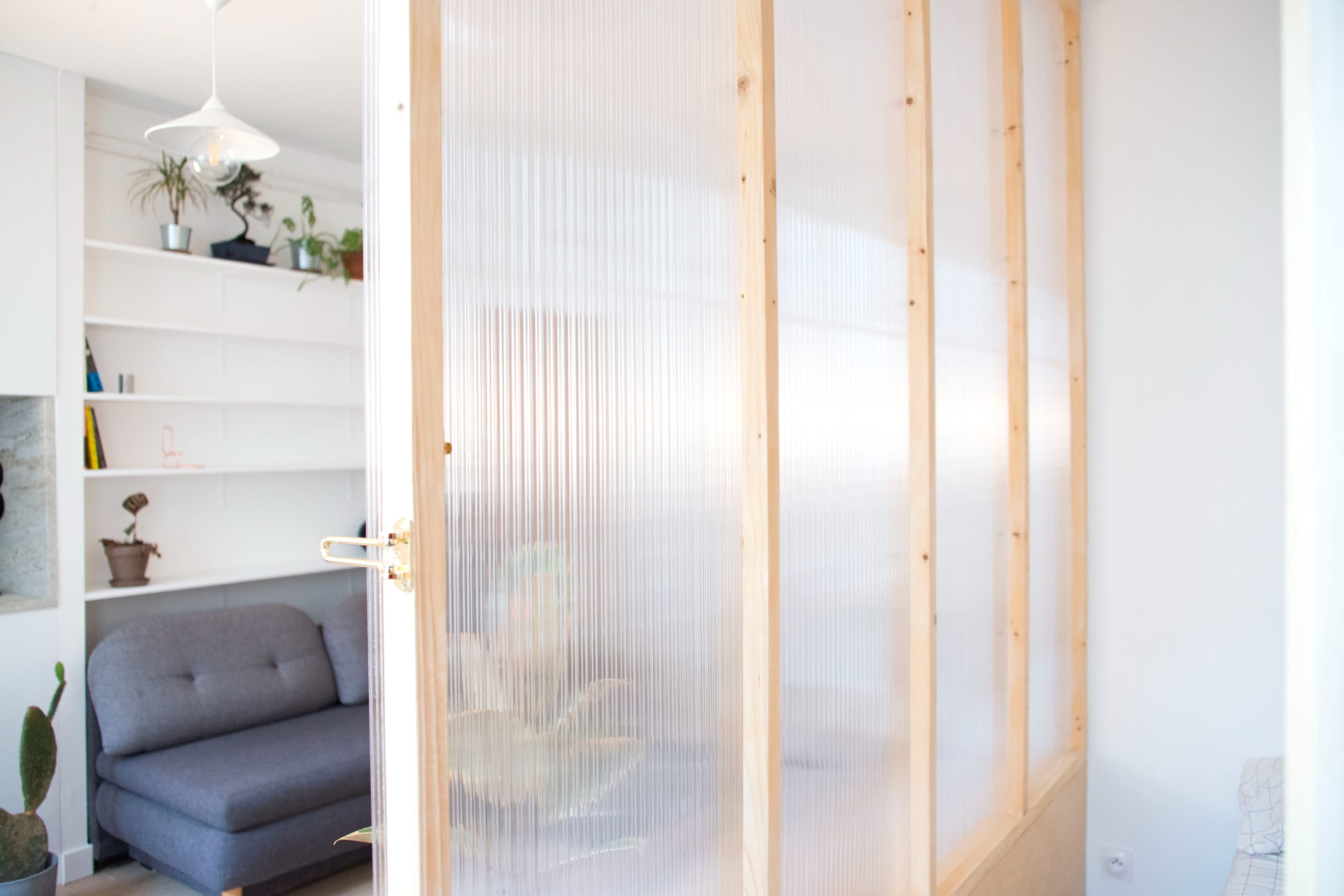 A translucent panel with wooden framing divides a living space, revealing a gray sofa and decorative plants in the background.
