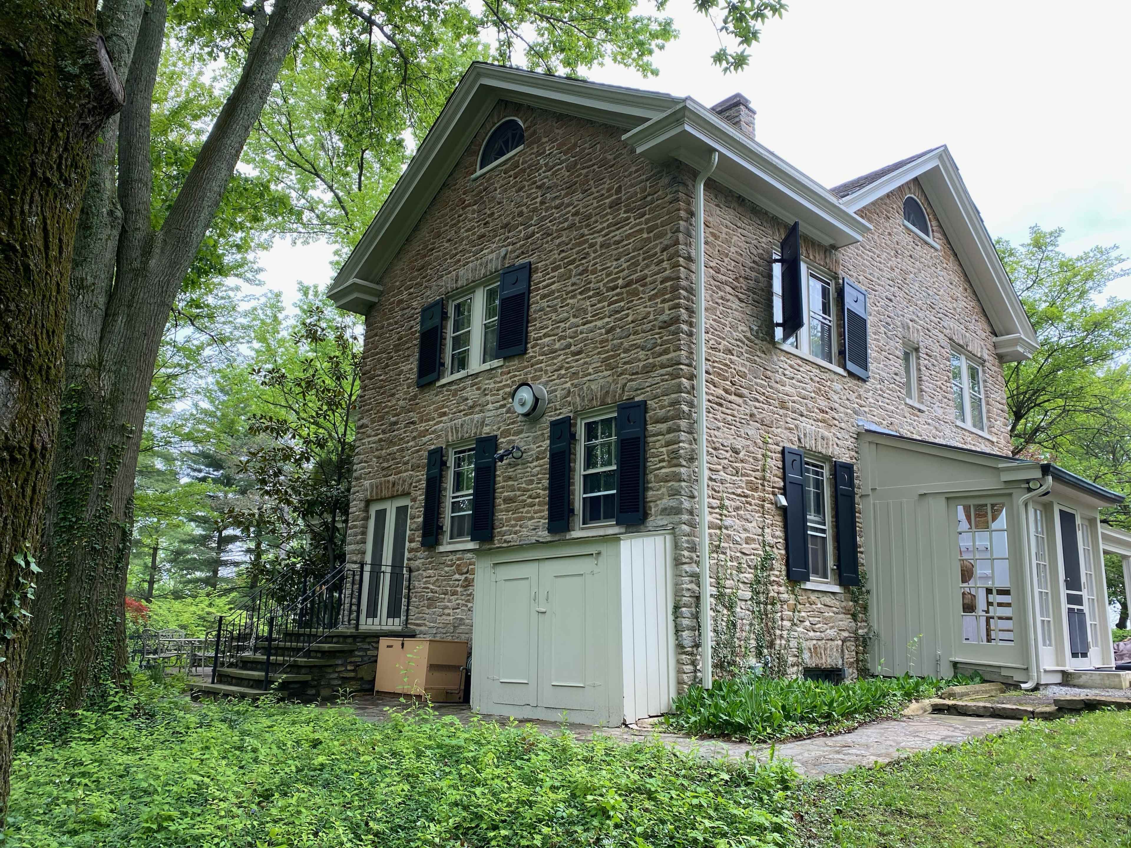 A two-story stone house with black shutters and a white addition, surrounded by greenery and trees.