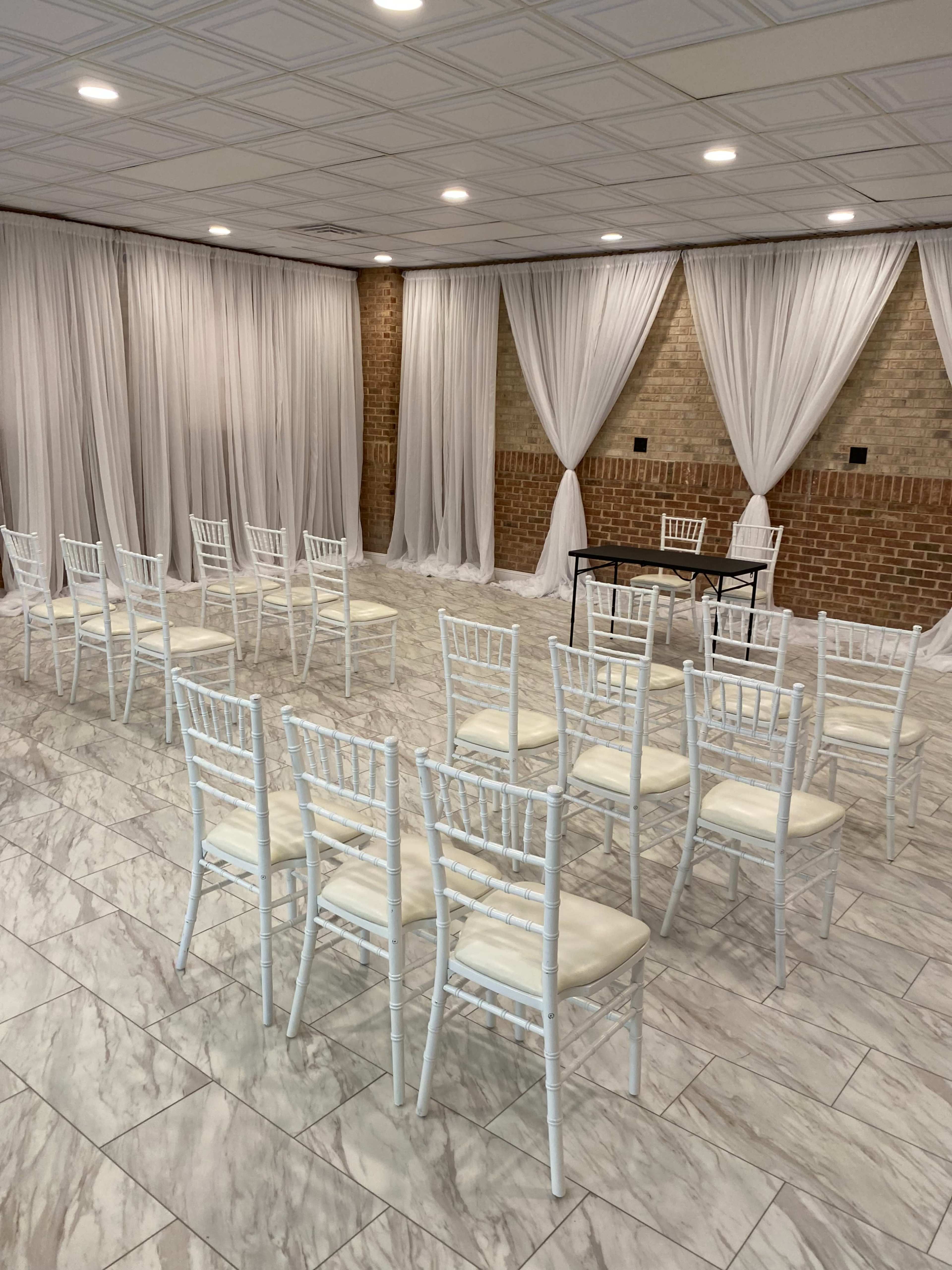 A spacious room features white chiavari chairs arranged in rows facing a small table, with sheer drapes hanging on the walls and a brick accent behind.
