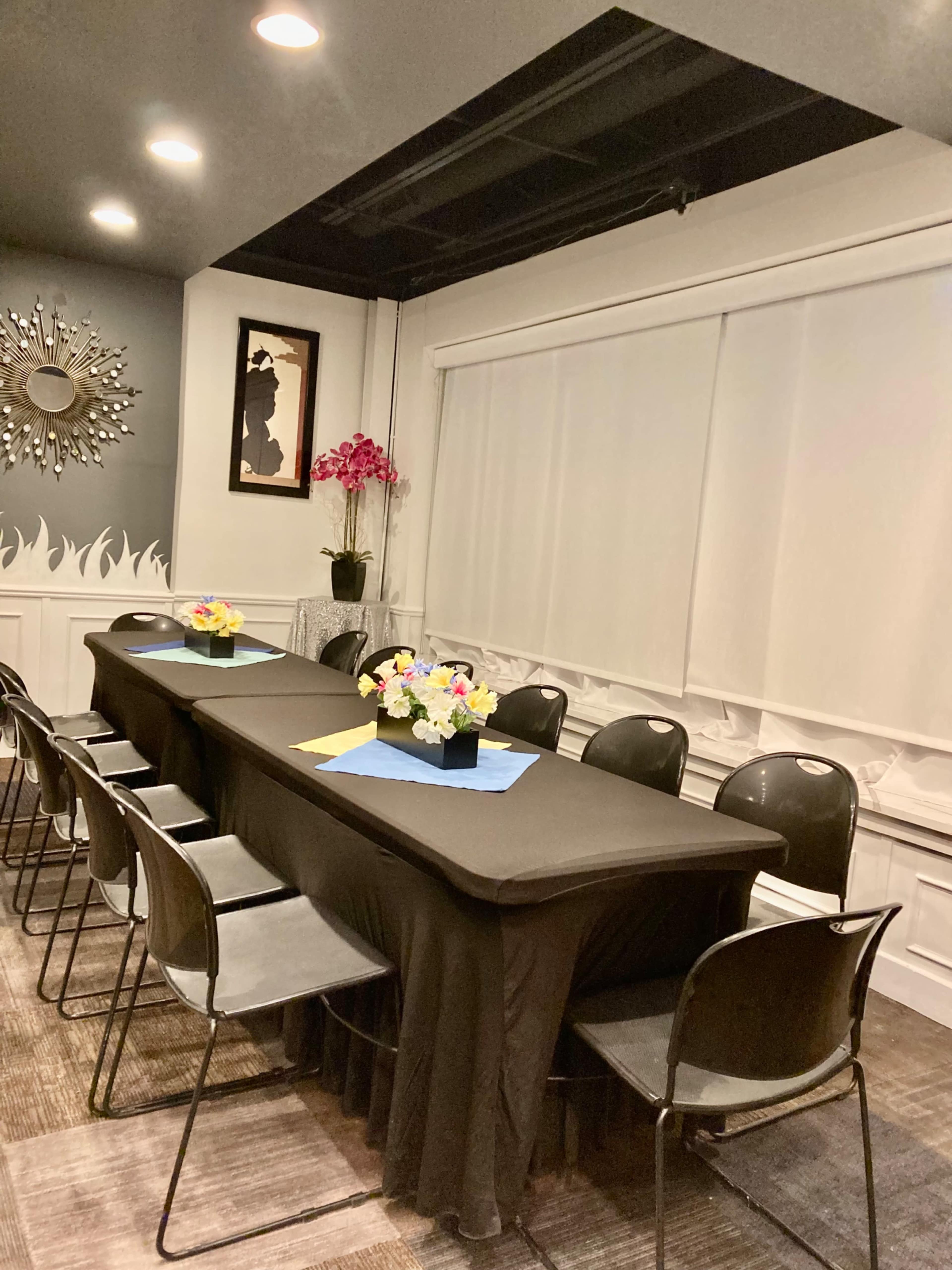 A long black table is set with flower centerpieces in a neatly arranged meeting room with black chairs and soft lighting.