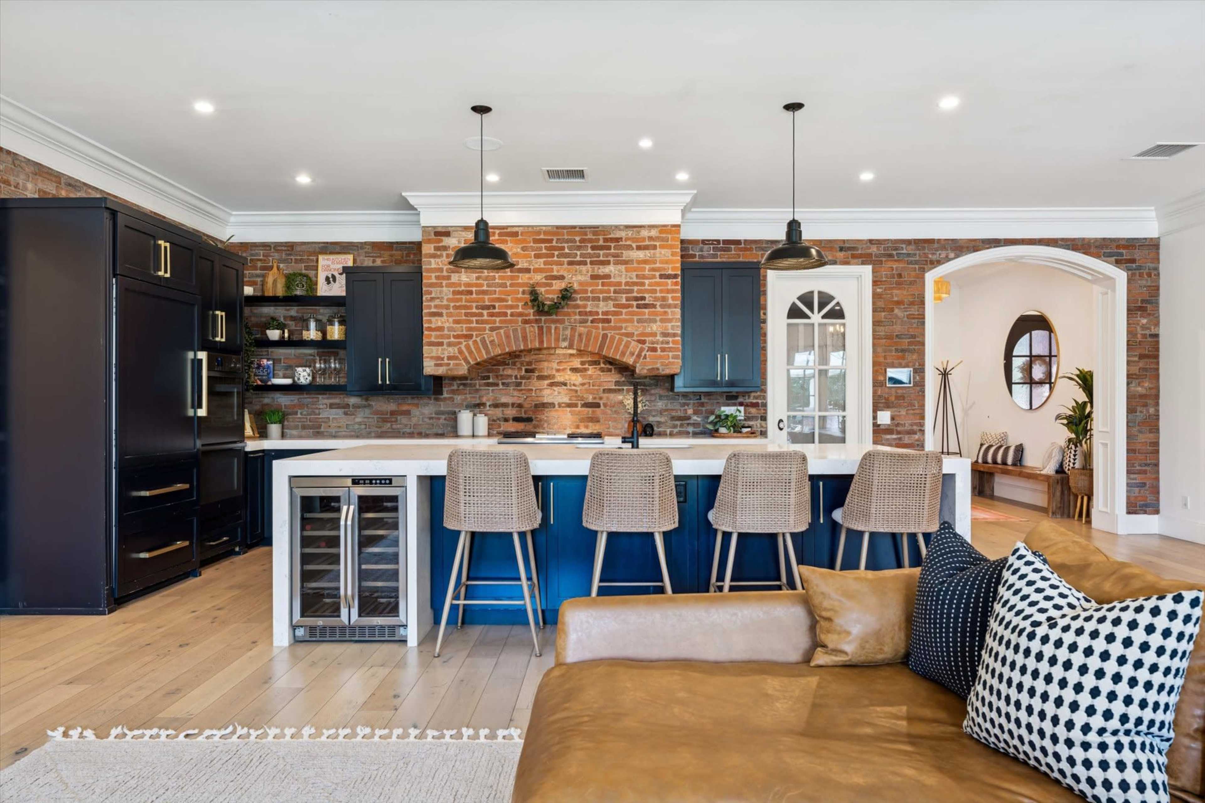 The image shows a modern kitchen featuring a brick wall, dark cabinetry, and a large island with bar stools.