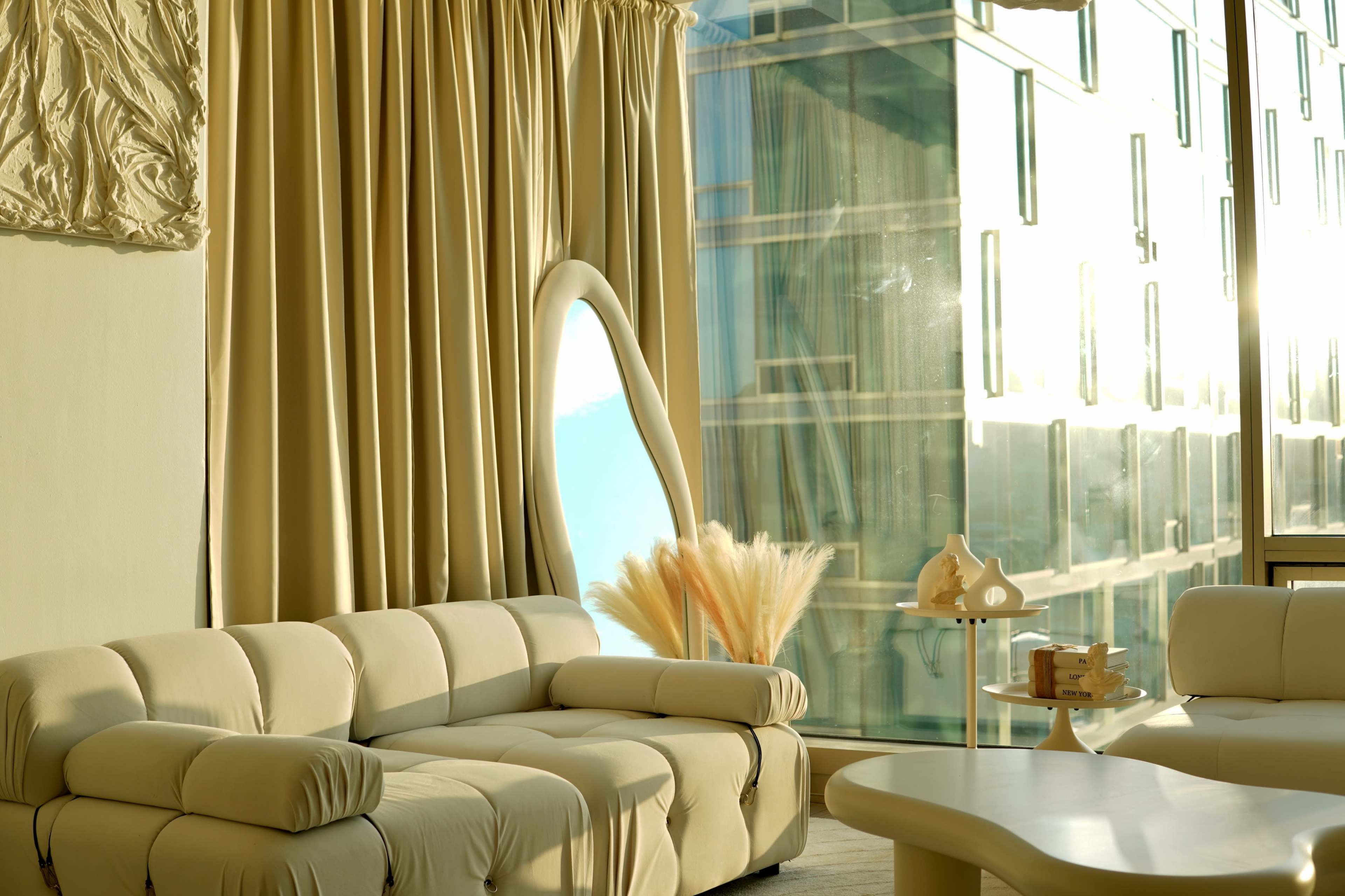A modern, minimalist living room with a curved sofa, large drapes, and a decorative arrangement of dried plants by a large window.
