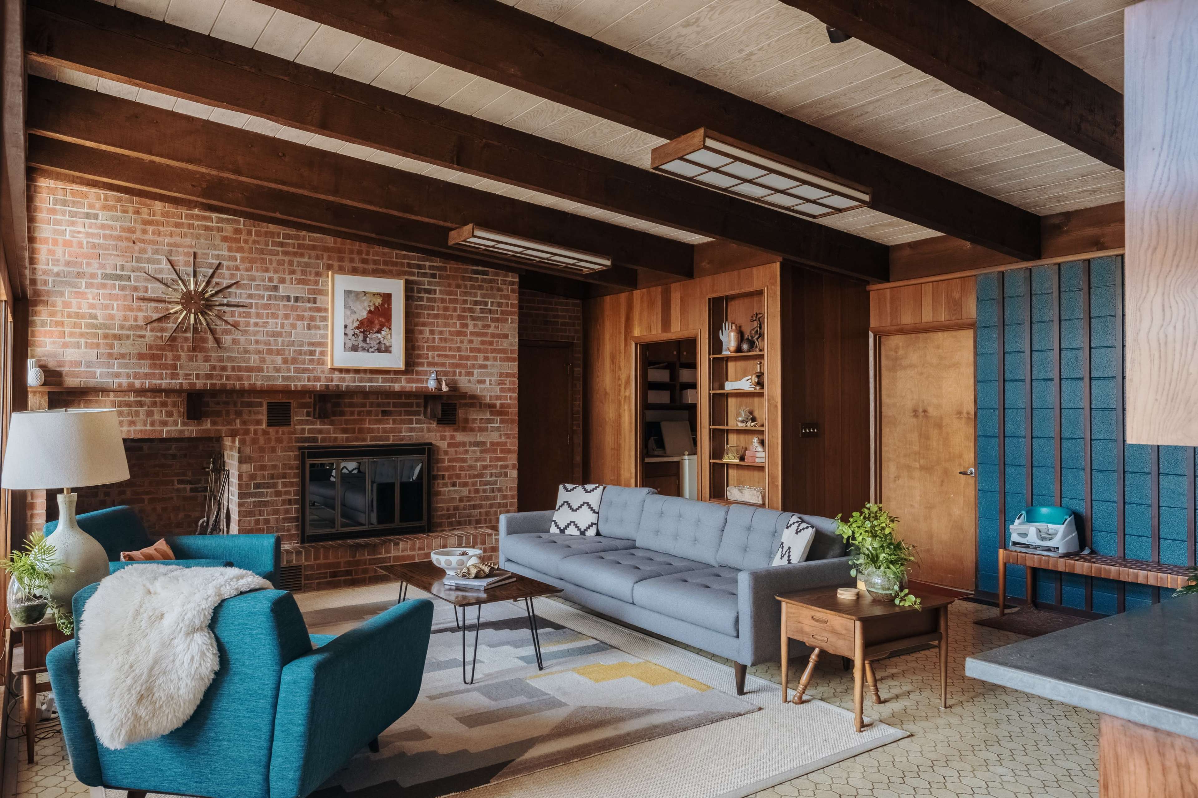 A modern living room features a gray sofa, teal armchairs, a wooden coffee table, and a brick wall with a fireplace, accented by wooden beams overhead.