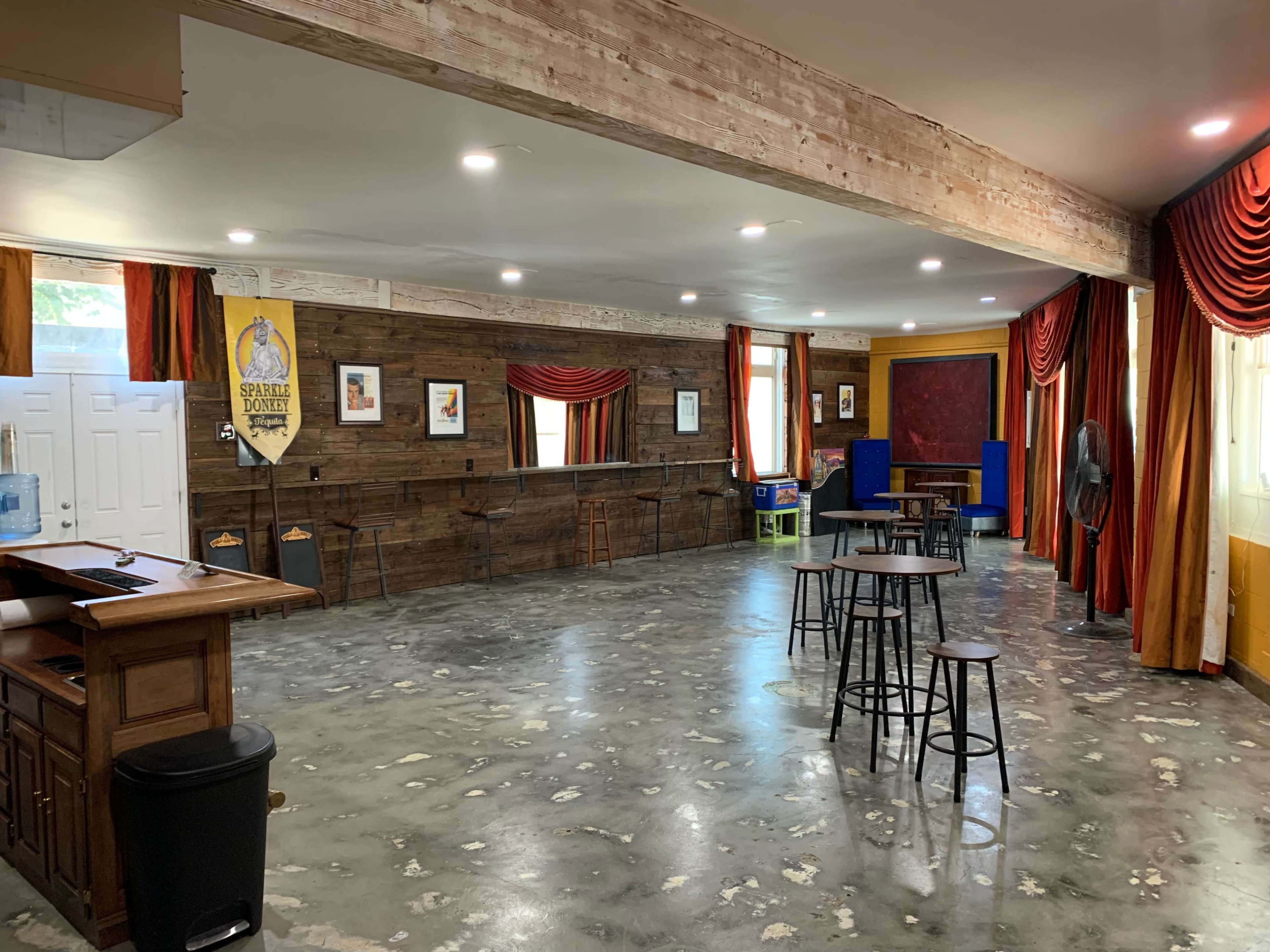 A spacious room with wooden walls, a polished concrete floor, various tables and chairs, and red drapes framing windows.