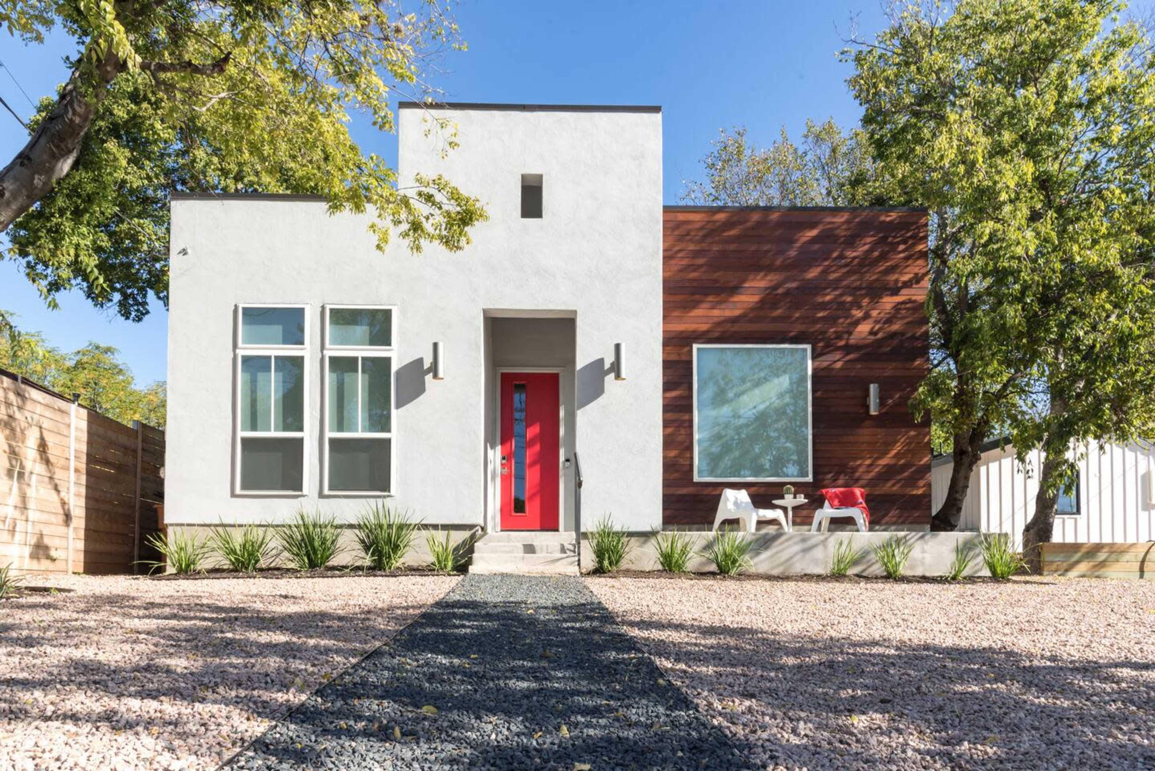 A modern house with a red door, framed by large windows and accented with wooden panels, is surrounded by a landscaped yard and a gravel pathway.